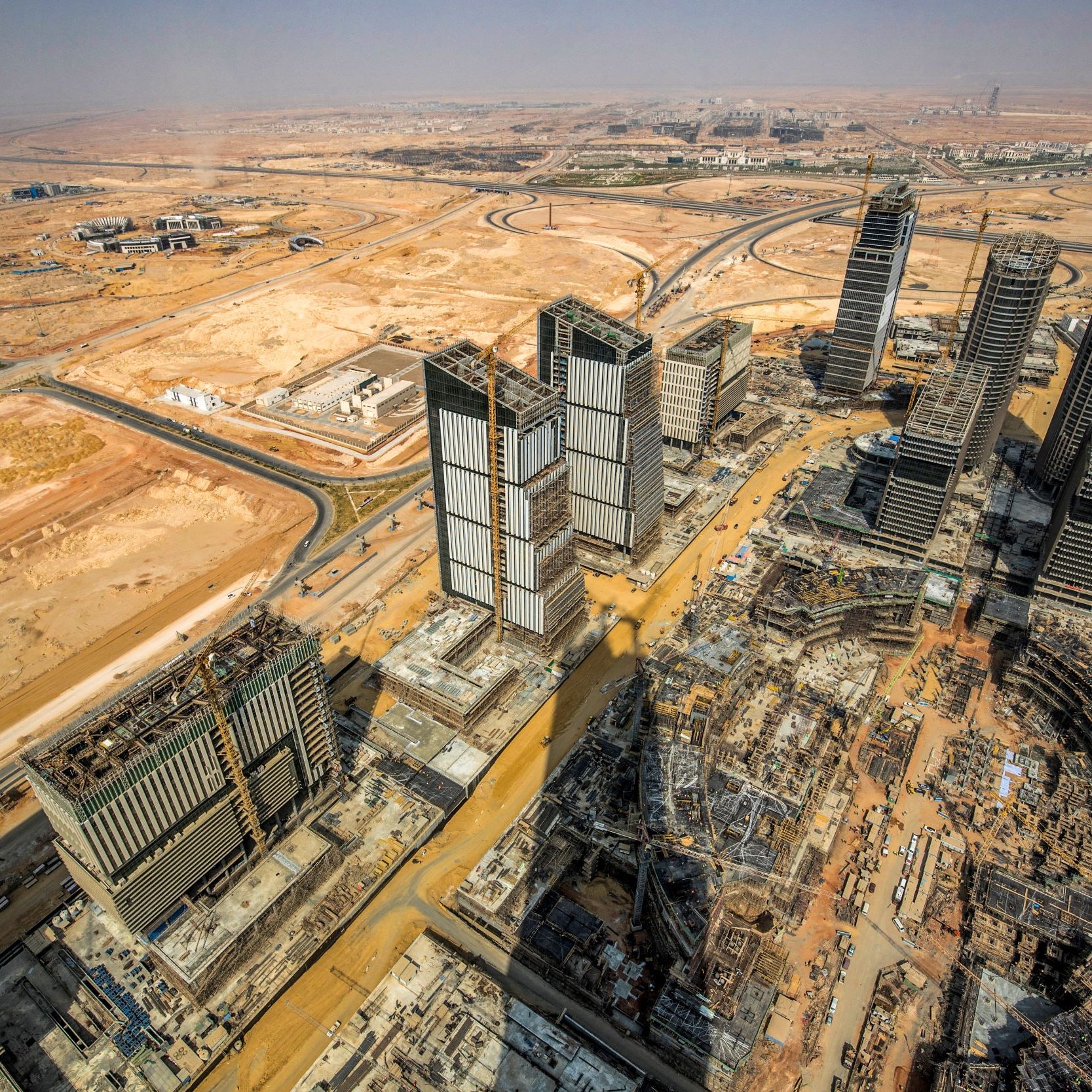 L'image montre une vue aérienne d'une région urbaine en développement, probablement dans un environnement désertique. On peut voir plusieurs gratte-ciels en construction, avec des structures modernes et des tours au milieu. Le paysage environnant est principalement désertique, avec peu de verdure, et des routes qui serpentent à travers la zone. La perspective donne une impression de grandeur et de modernité, tout en mettant en évidence les travaux d'infrastructure en cours.