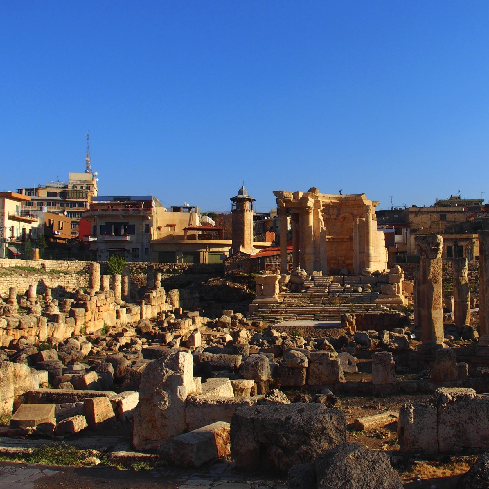 این تصویر نمایی از یک مکان باستانی را نشان می‌دهد که شامل خرابه‌های ستون‌ها و دیوارهای قدیمی است. در پس‌زمینه، چند ساختمان مدرن‌تر دیده می‌شود که نشان‌دهنده ترکیبی از تاریخ با معماری معاصر است. آسمان آبی و صاف است و نور خورشید بر روی سنگ‌ها می‌تابد که حس زیبایی و آرامش را به تصویر می‌بخشد. اجزای باستانی این مکان نشان‌دهنده تاریخ غنی و فرهنگی آنجا هستند.