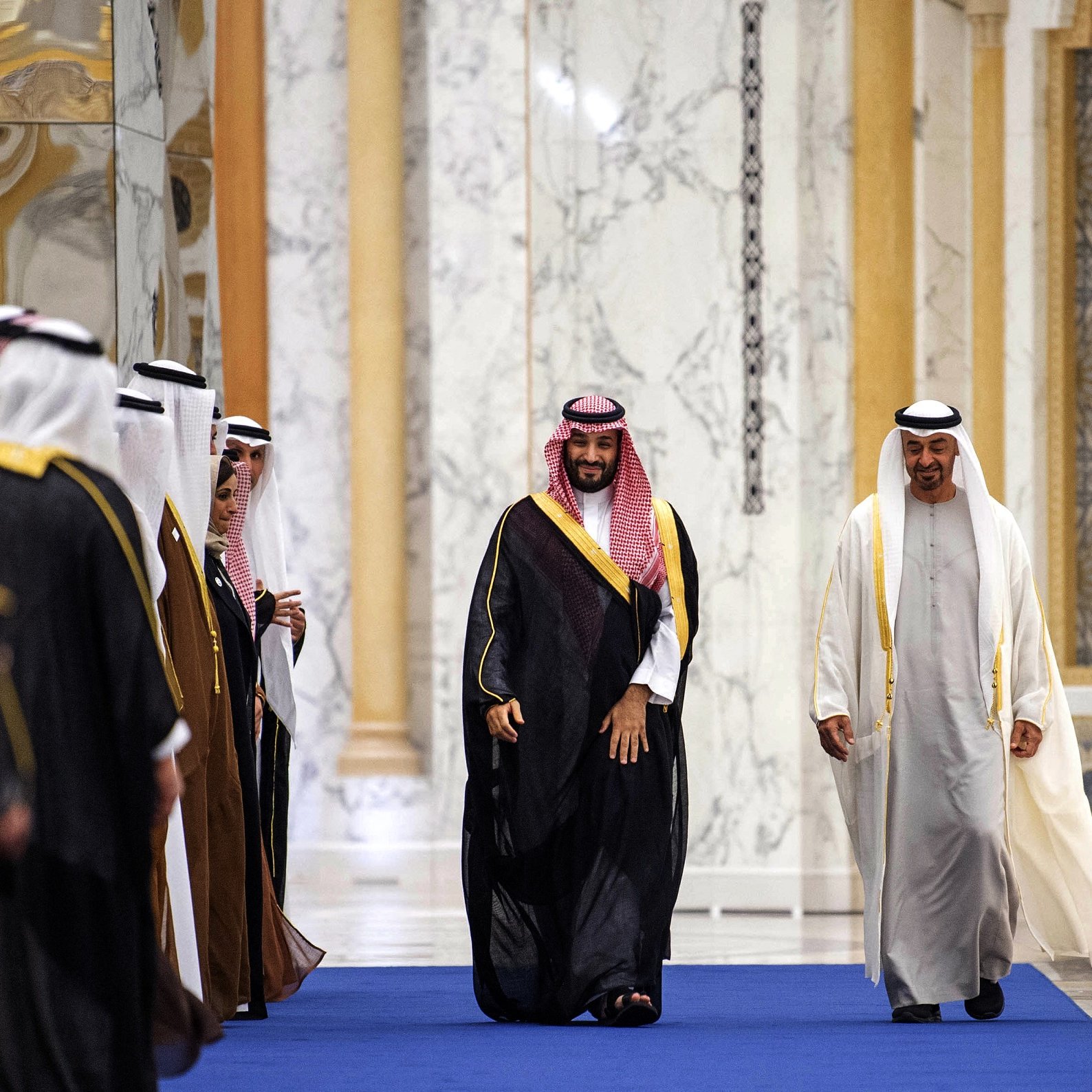 در این تصویر، گروهی از مردان به صورت رسمی در یک محیط داخل ساختمان قرار دارند. برخی از آنها لباس‌های سنتی عربی به تن دارند و در حال خوش‌آمدگویی یا استقبال از یک نفر دیگر هستند که به آرامی در حال عبور از میان آنهاست. شخصی که در مرکز تصویر قرار دارد، لباسی متفاوت و برجسته‌تر دارد و به نظر می‌رسد نقش مهم‌تری در این مراسم بازی می‌کند. محیط با طراحی مجلل و دیوارهای سنگی تزئین شده است.