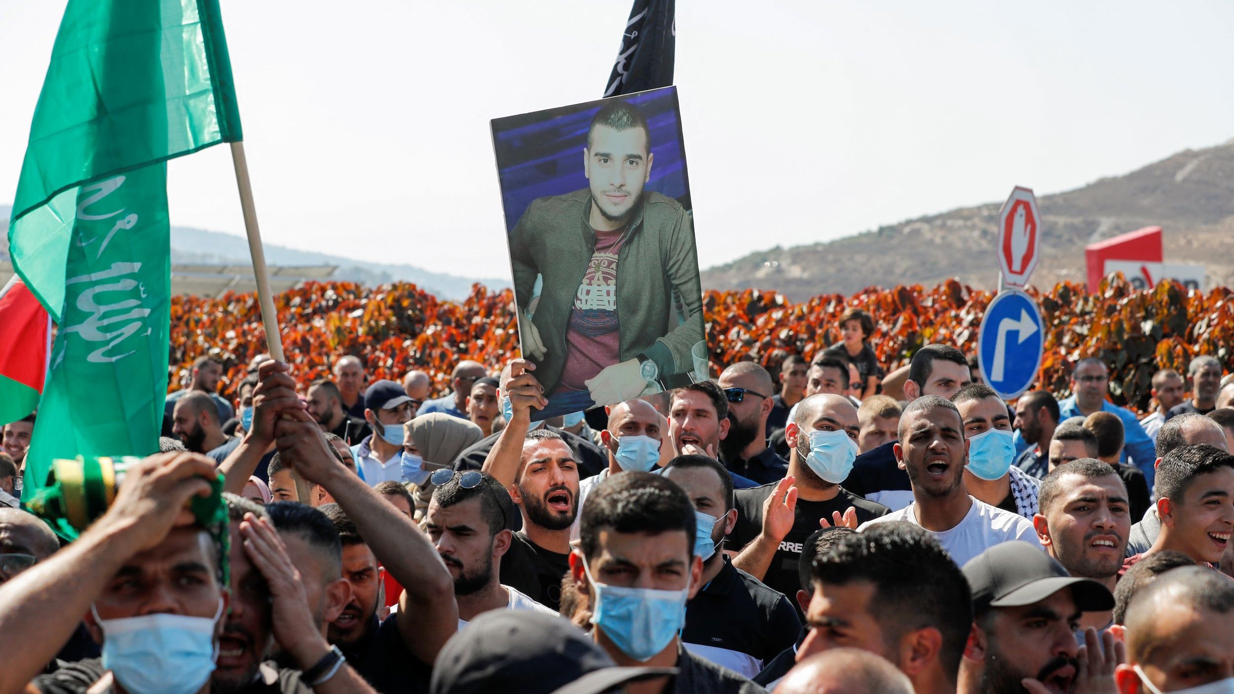 الصورة تُظهر مجموعة من المتظاهرين في تجمع حاشد، يحمل بعضهم لافتات وأعلام. وسط الحشد، يوجد شخص يحمل صورة لرجلٍ يبدو أنه شخصية مهمة أو رمزية بالنسبة لهم. يرتدي الكثير من المتظاهرين الكمامات، ويبدو أن المظاهرة تتم في منطقة مفتوحة، مع خلفية تضم تلالًا وأشجار. تُعبر الأجواء عن الحماس والتجمع الجماهيري.