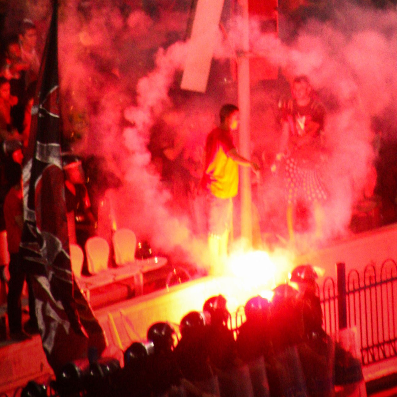 L'image montre une scène de manifestation avec une atmosphère intense. On peut voir des fumigènes rouges créant une nuée de fumée, ainsi que des personnes rassemblées autour d'une structure ou d'un symbole marquant, probablement dans un contexte de protestation. Au premier plan, une file de policiers en uniformes semble faire face aux manifestants. L'ensemble de la scène transmet une impression de tension et d'engagement émotionnel.