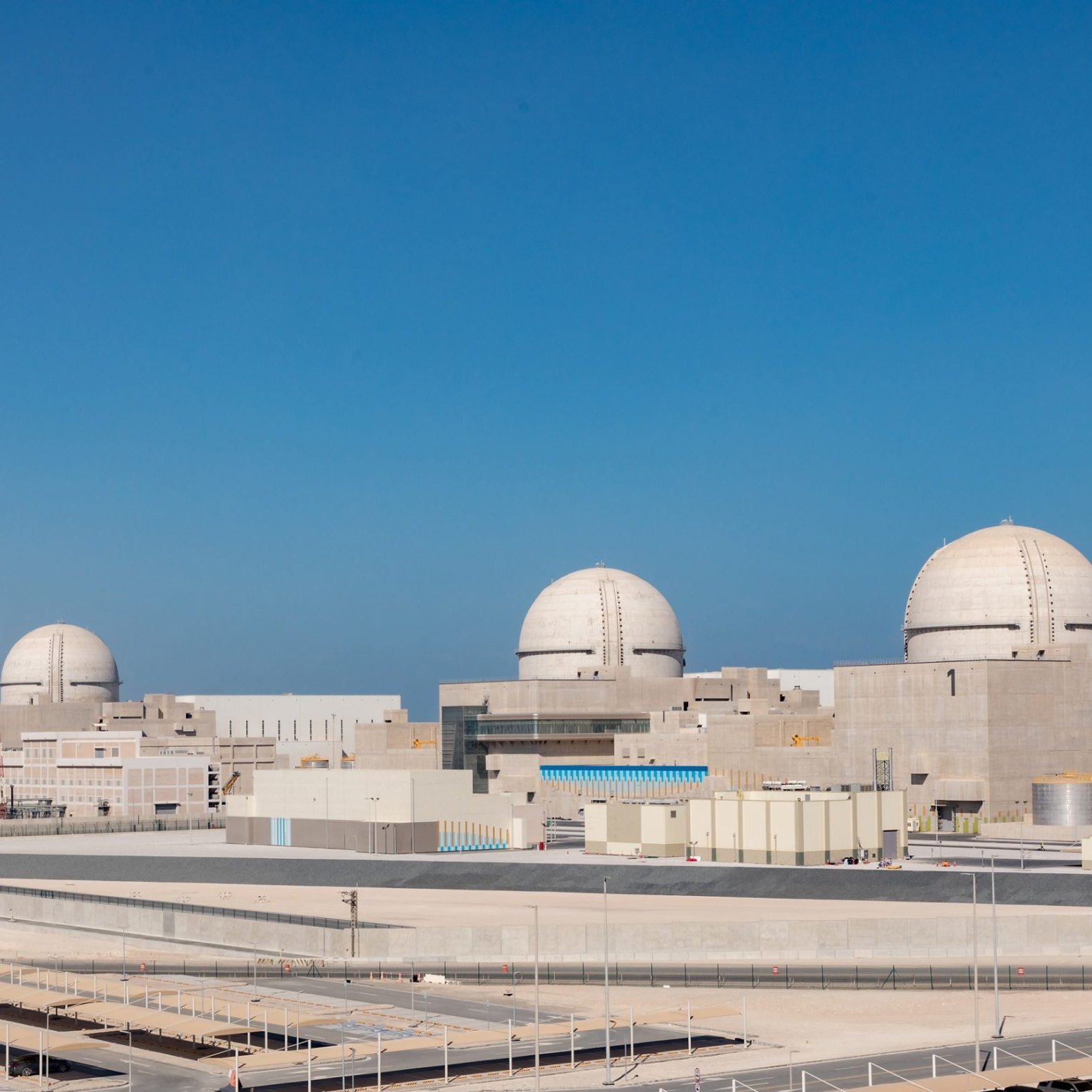 این تصویر نشان‌دهنده یک نیروگاه هسته‌ای است که دارای چهار گنبد بزرگ در بالای ساختمان‌ها می‌باشد. این گنبدها معمولاً به عنوان محافظ برای راکتورهای هسته‌ای استفاده می‌شوند. پس‌زمینه آسمان آبی و صاف است و ساختارهای بتنی نیروگاه در جلوی تصویر قرار دارند. به نظر می‌رسد این تاسیسات مدرن و صنعتی هستند و در یک منطقه سرسبز یا شهری قرار دارند.