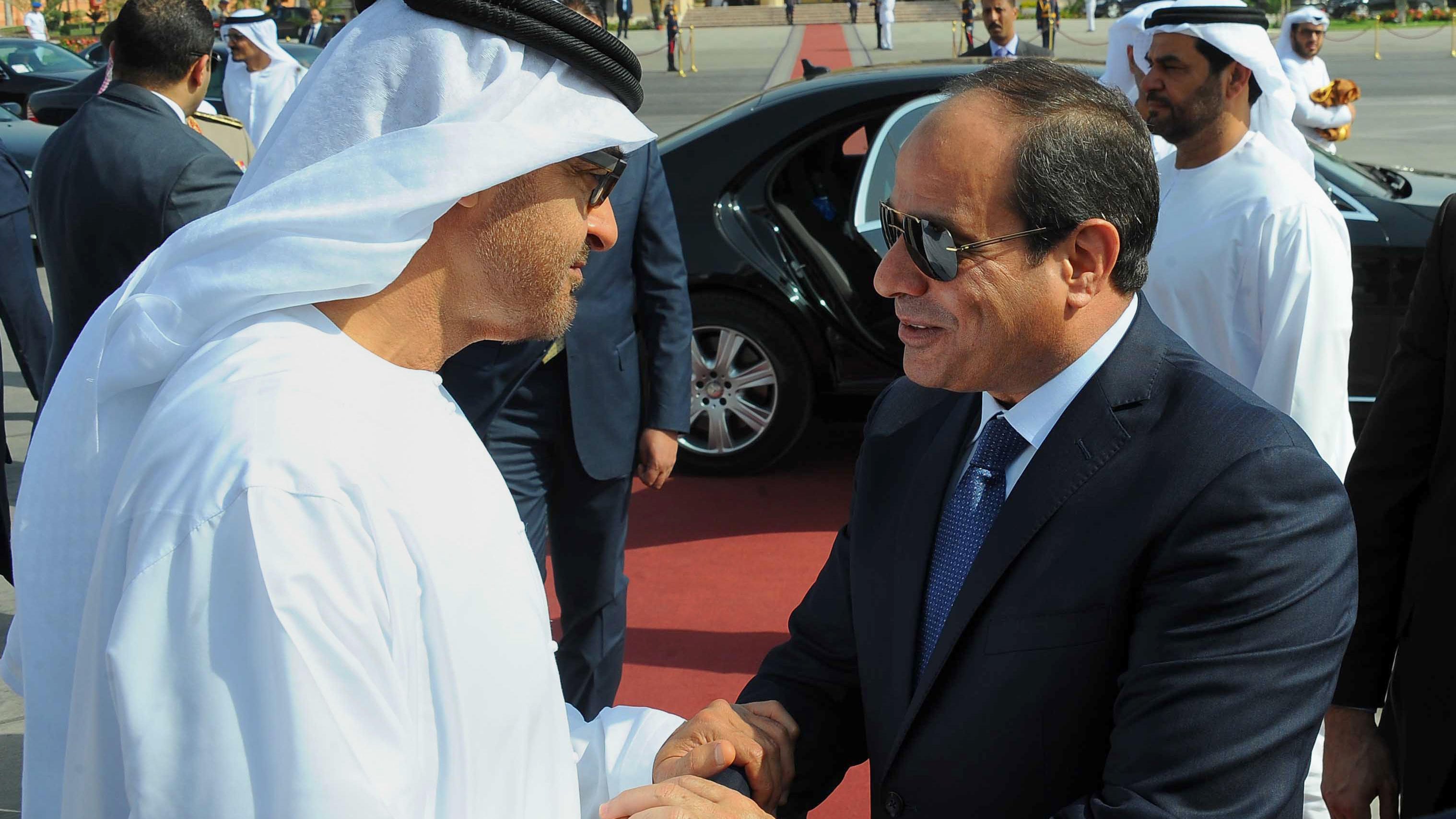 L'image montre une rencontre entre deux dirigeants, probablement en train de se saluer avec une poignée de main. Les individus sont vêtus de vêtements traditionnels, comme des kandouras et des keffiehs pour l'un d'eux. On peut également voir d'autres personnes en arrière-plan, ainsi qu'une voiture haut de gamme, suggérant un événement officiel ou une cérémonie. L'ambiance semble cordiale et respectueuse.