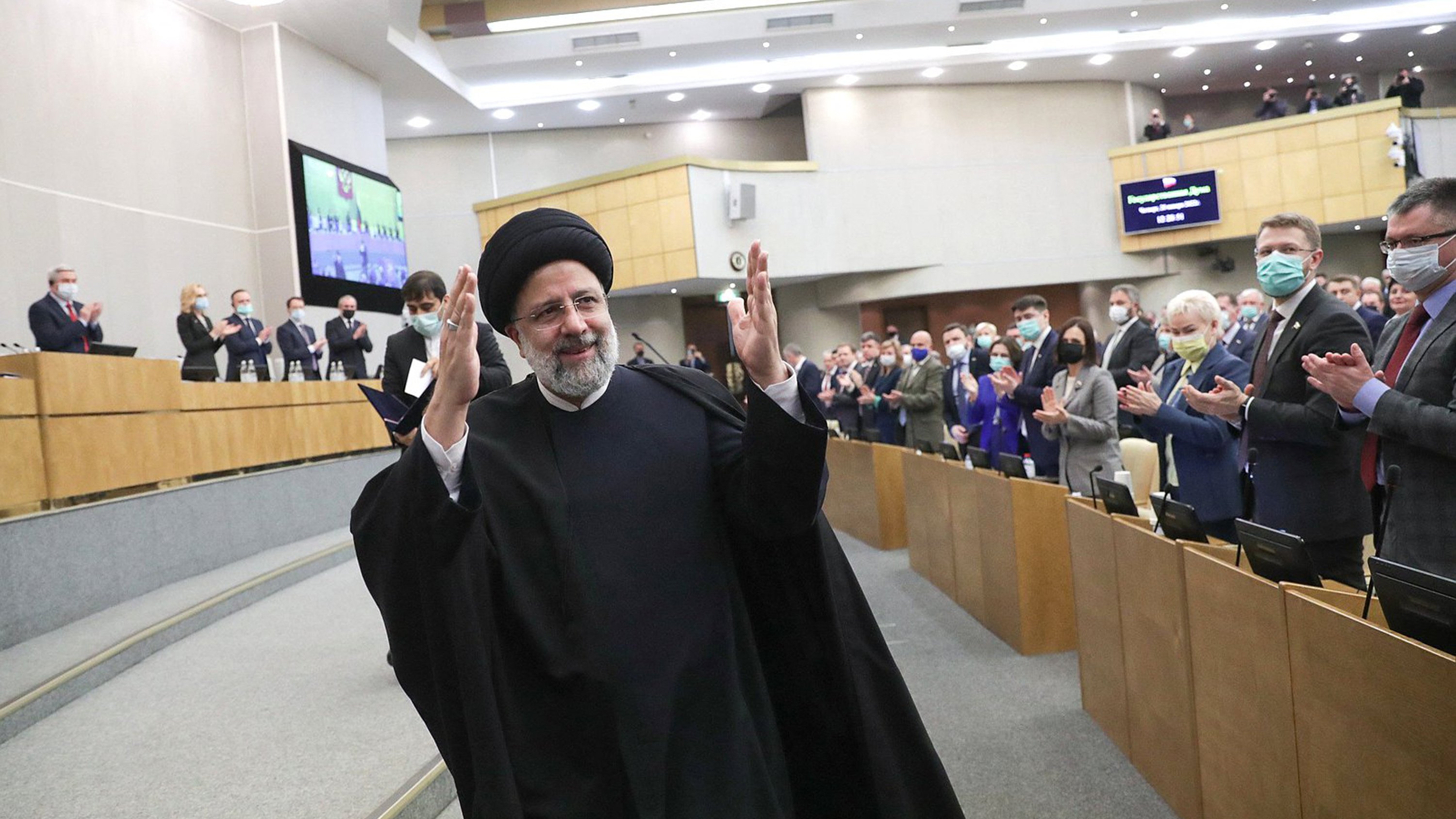 L'image montre un homme en habit traditionnel, portant un turban noir, qui semble être en train de saluer un public. Il se tient au centre d'une salle où plusieurs personnes applaudissent. L'environnement est formel, typique d'un événement politique ou d'une réunion officielle, avec des sièges et des écrans visibles en arrière-plan. Les participants portent également des masques de protection, indiquant une attention à la santé publique.