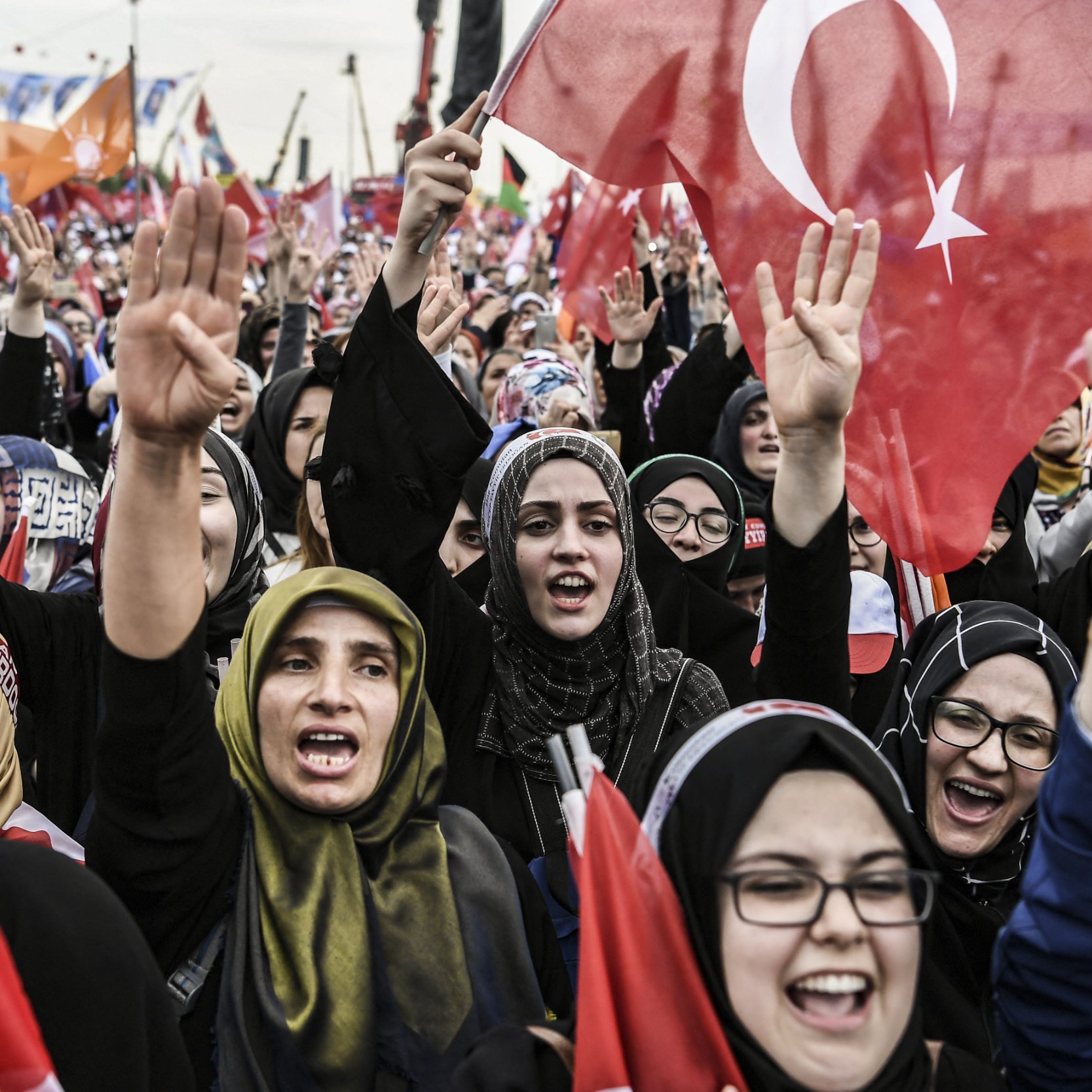 الصورة تظهر حشودًا من النساء في تظاهرة أو فعالية جماهيرية. العديد من النساء يحملن الأعلام التركية ويعبرن عن حماسهن من خلال رفع الأيادي. يبدو أنهن يرتدين ملابس تقليدية أو حجاب، مما يعكس تنوع أساليب التعبير عن الهوية. الجو العام يظهر حيوية وشغفًا، مع وجود لافتات وأعلام في الخلفية تدل على رسالة أو هدف مشترك.