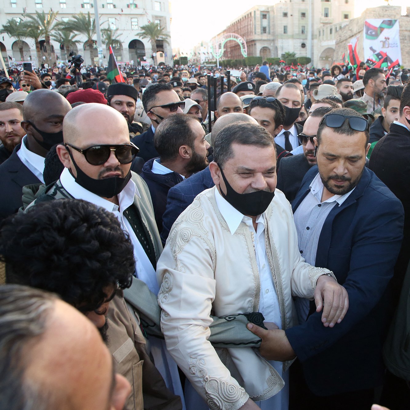 در این تصویر، جمعیتی از مردم در یک رویداد یا مراسم خیابانی دیده می‌شود. در مرکز تصویر، مردی در لباس سنتی روشن در حال احاطه شدن توسط چند نفر دیگر است که بعضی از آن‌ها ماسک زده‌اند. به نظر می‌رسد که او مورد توجه و استقبال زیادی قرار گرفته و در حال تعامل با دیگران است. فضای اطراف پر از افرادی است که در حال تماشا یا شرکت در این رویداد هستند و پرچم‌هایی در دست دارند. در زمینه، ساختمان‌ها و فضاهای عمومی نیز مشخص است.