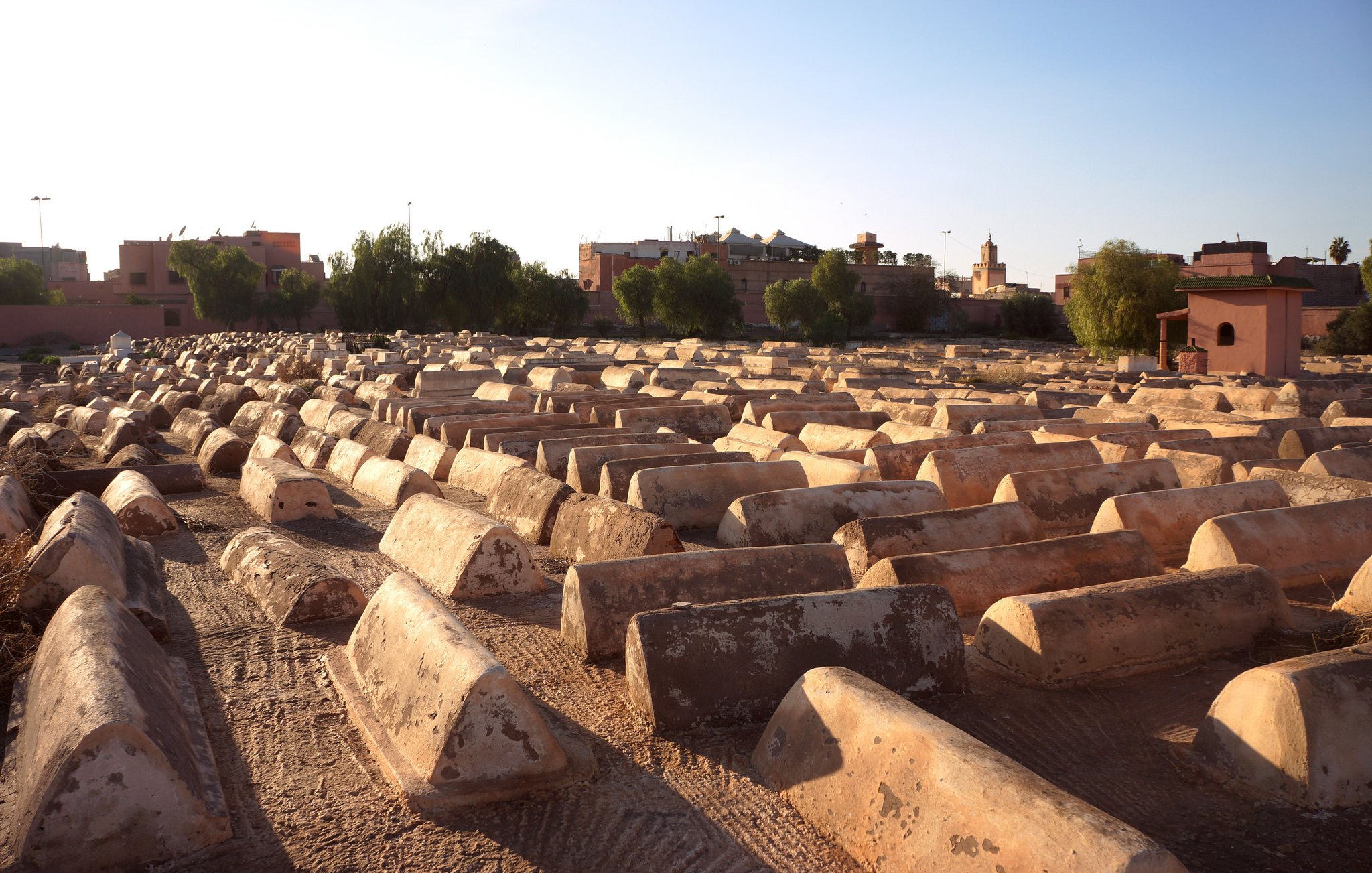 L'image montre un cimetière où l'on peut voir des tombes en forme de tombeaux, étalées sur un terrain sec. Les structures sont de couleur beige ou terre cuite et semblent être en béton, avec une surface usée par le temps. En arrière-plan, on aperçoit des bâtiments et des arbres, indiquant que le cimetière est situé près d'une ville. La lumière du soleil crée une atmosphère calme et paisible, tandis que le ciel est dégagé, laissant entrevoir un beau jour.