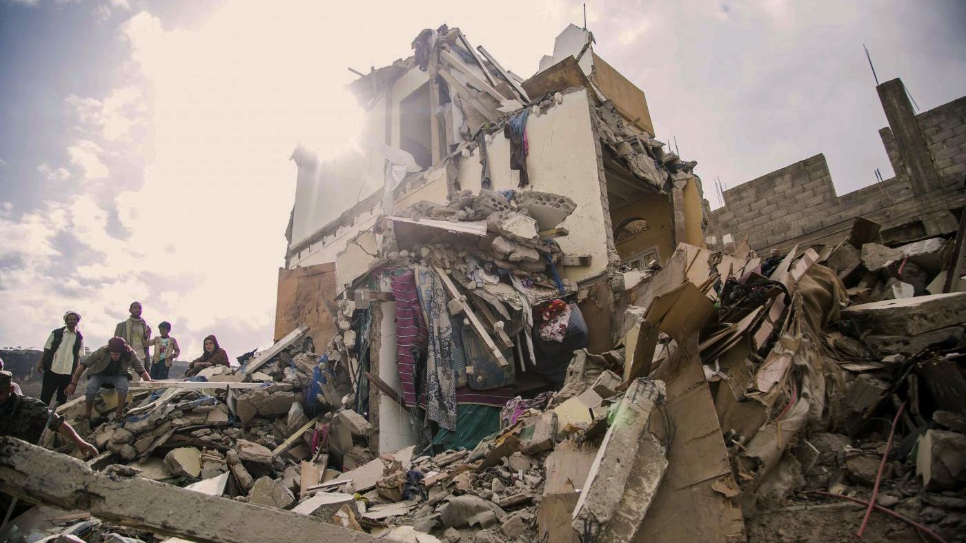 L'image montre un bâtiment effondré, témoignant des dégâts causés par un tremblement de terre ou une catastrophe similaire. Les débris sont éparpillés autour, et on peut voir des personnes s'approchant des ruines. La lumière du soleil perçoit à travers les restes du bâtiment, ajoutant une atmosphère dramatique à la scène de destruction. Les couleurs sont ternes et le paysage donne une impression de désolation et de tristesse face à la situation.