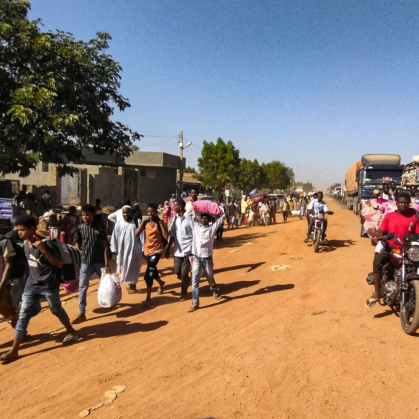 L'image montre une scène de rue animée dans une zone urbaine. Sur le côté gauche, on voit un arbre et des bâtiments, tandis que des gens marchent le long de la route en terre. Parmi eux, certains portent des sacs ou des charges sur la tête. Des véhicules, y compris des camions et des motos, circulent sur la route, créant une ambiance dynamique. L'éclairage semble être en plein jour, suggérant une activité quotidienne typique dans cette communauté.