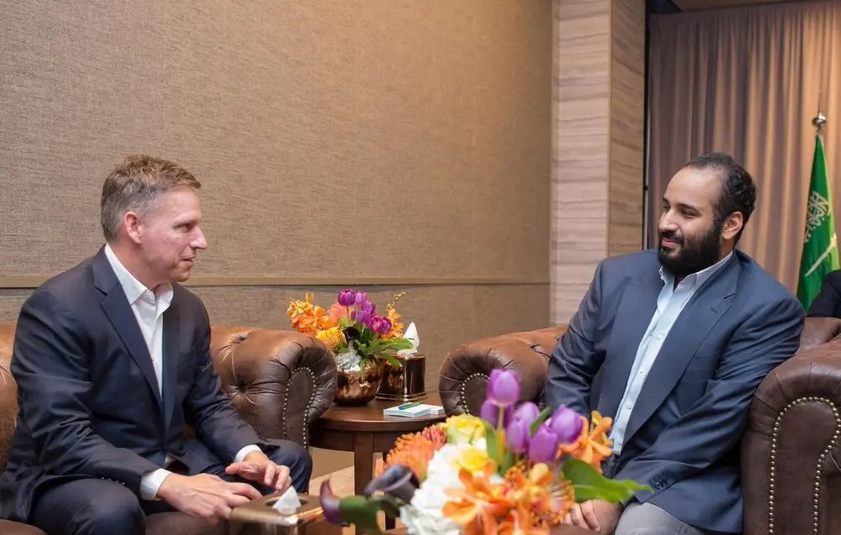 دو مرد در یک اتاق مرتب نشسته‌اند. یکی با کتی تیره و دیگری با پیراهن روشن. گل‌های رنگارنگ روی میز است.