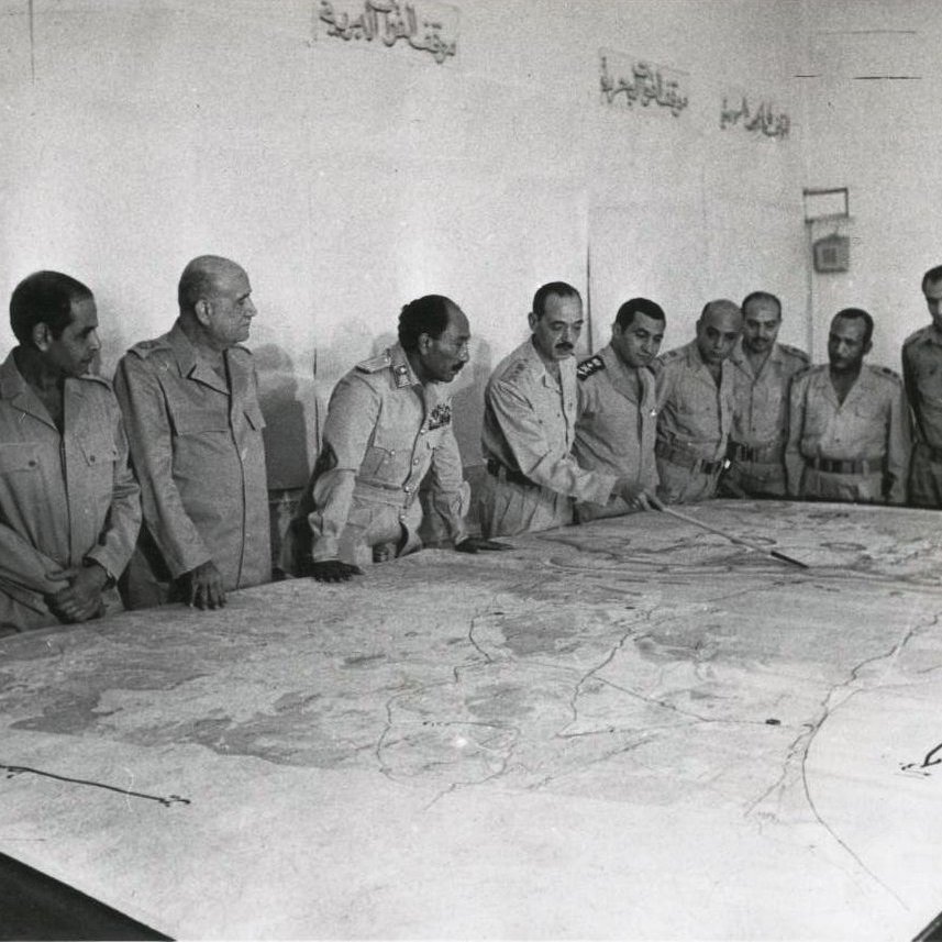 L'image montre un groupe d'hommes en uniformes militaires se tenant autour d'une grande carte posée sur une table. Ils semblent concentrés et discutent de la carte, qui est détaillée avec des lignes et des zones géographiques. L'environnement est clair et sobre, avec des murs nus. L'atmosphère semble sérieuse, probablement en rapport avec des stratégies militaires.