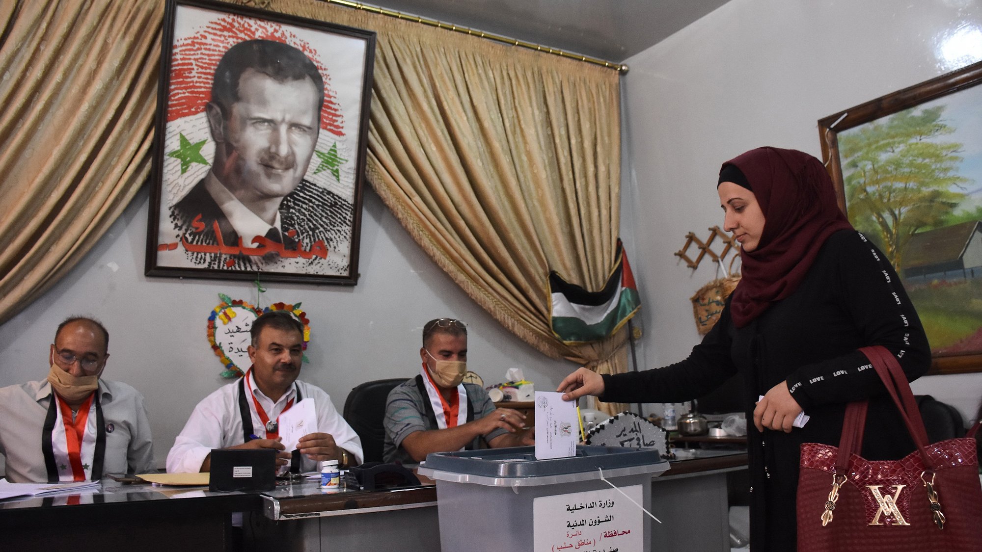 تظهر الصورة امرأة ترتدي عباءة وحجابًا تضع ورقة اقتراع في صندوق الانتخاب. خلفها، يوجد لوحة كبيرة تحمل صورة شخصية معروفة. يجلس ثلاثة رجال خلف طاولة، حيث يبدو عليهم الاستعداد لمراقبة العملية الانتخابية. المكان يبدو رسميًا، مع ستائر وخلفية تشير إلى الطابع المحلي.