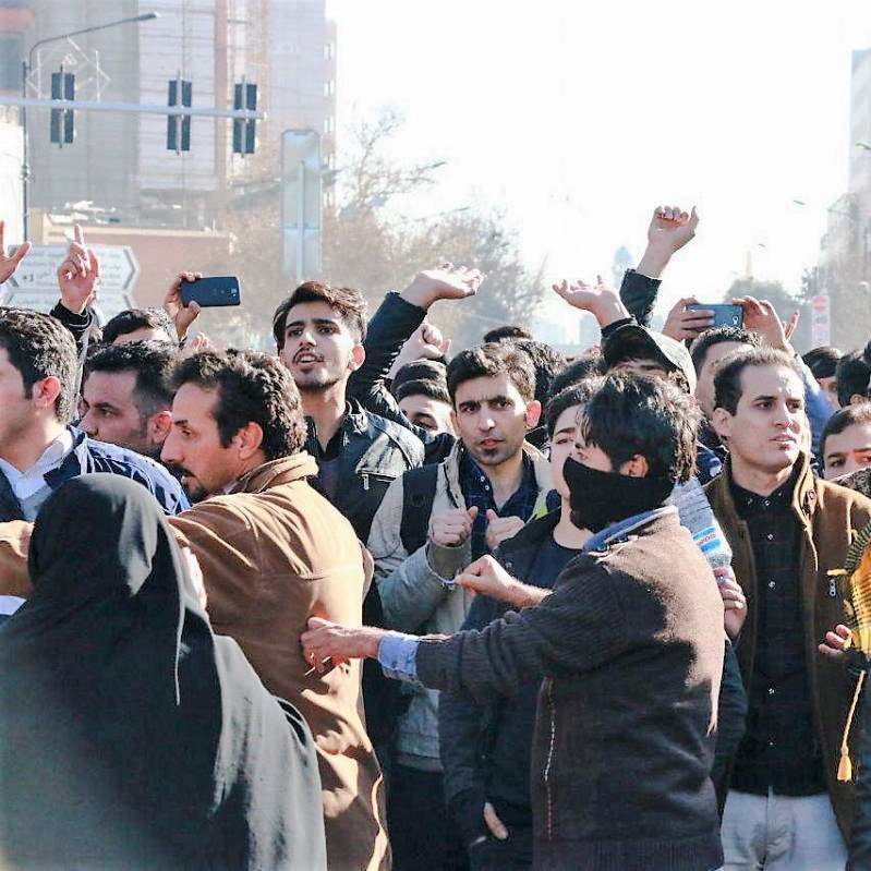 در این تصویر، گروهی از مردم در یک جمعیت انبوه دیده می‌شوند که به نظر می‌رسد در حال برگزاری یک تجمع یا اعتراض هستند. برخی از افراد دست‌های خود را بالا برده‌اند و دیگران به سمت دوربین نگاه می‌کنند. تعدادی از افراد لباس‌های معمولی بر تن دارند و در پس‌زمینه، ساختمان‌ها و چراغ‌های راهنمایی قابل مشاهده است. حالت چهره‌ها نشان‌دهنده‌ی هیجان یا تمایل به ابراز نظر است. برخی از افراد نیز با پوشش صورت دیده می‌شوند.