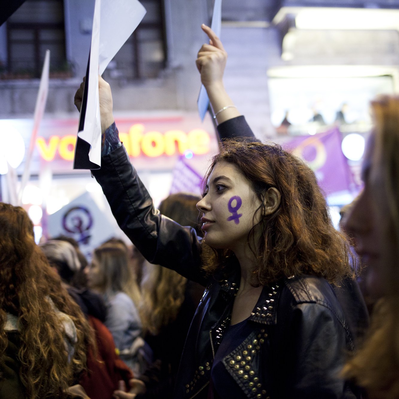 الصورة تُظهر مجموعة من المتظاهرين في تظاهرة تحتفل بحقوق المرأة. في المقدمة توجد امرأة شابة تحمل ورقة وتُظهر رمز أنثوي مرسوم على وجهها. ترتدي سترة جلدية وتبدو مفعمة بالحماس. في الخلفية، يمكن رؤية أعلام ومستندات تحمل شعارات تخص حركة النساء، مع أجواء من الحماس والتضامن بين المشاركين.