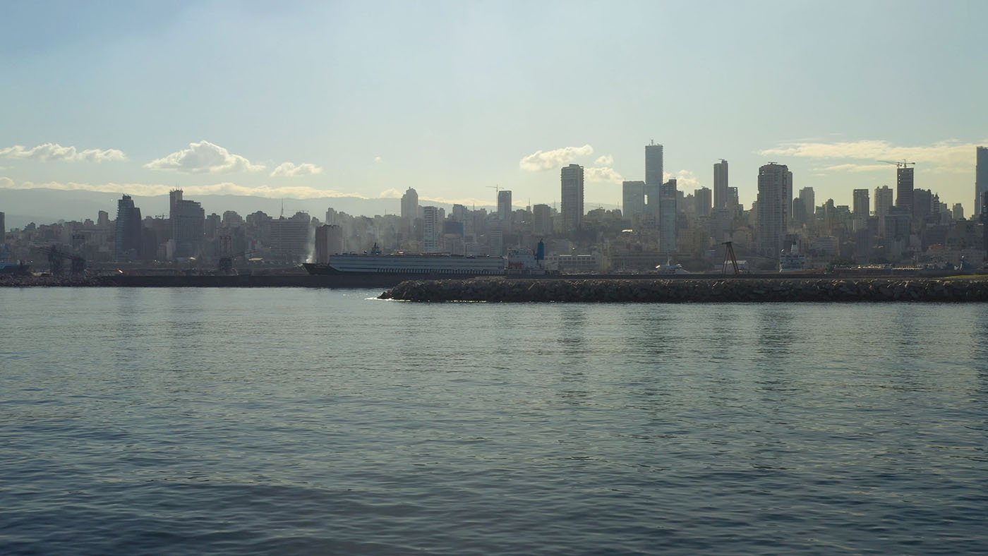 L'image montre un paysage urbain d'une ville moderne en bord de mer. On peut voir des gratte-ciels s'élevant le long de la côte, reflétant la lumière du soleil sur l'eau calme de la mer. Au premier plan, il y a un port ou une jetée, et l'arrière-plan est rempli de bâtiments qui se dressent majestueusement. Le ciel est légèrement nuageux, ce qui crée une ambiance douce et mélancolique. L'ensemble donne une impression de tranquillité et de modernité.