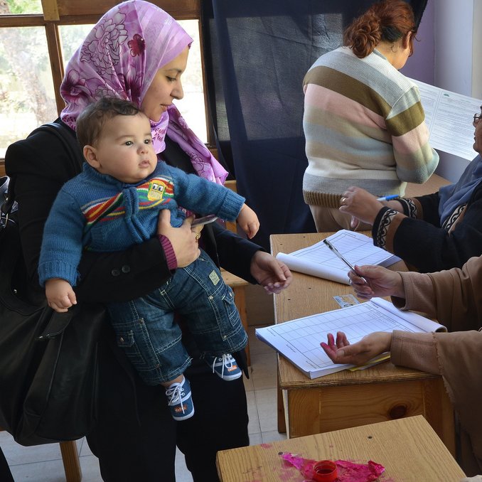 L'image montre une scène dans un environnement communautaire. On peut voir une femme portant un foulard qui tient un bébé dans ses bras, s'approchant d'une table où une autre femme, également couverte d'un foulard, est assise et prend des notes. À l'arrière-plan, d'autres femmes semblent occupées à diverses activités. L'ambiance semble être celle d'un événement organisé, possiblement une inscription ou une réunion. Les couleurs et les vêtements des femmes reflètent une culture riche et diversifiée.