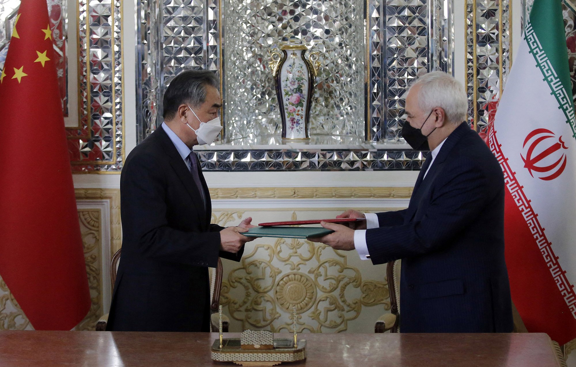 L'image montre deux hommes, apparemment des représentants de la Chine et de l'Iran, se tenant en face l'un de l'autre. Ils échangent des documents au cours d'une cérémonie formelle. On peut voir les drapeaux de la Chine et de l'Iran en arrière-plan. L'environnement est richement décoré, avec des motifs élaborés et un vase floral visible derrière eux. Les deux hommes portent des masques, ce qui pourrait suggérer que la photo a été prise pendant la pandémie.