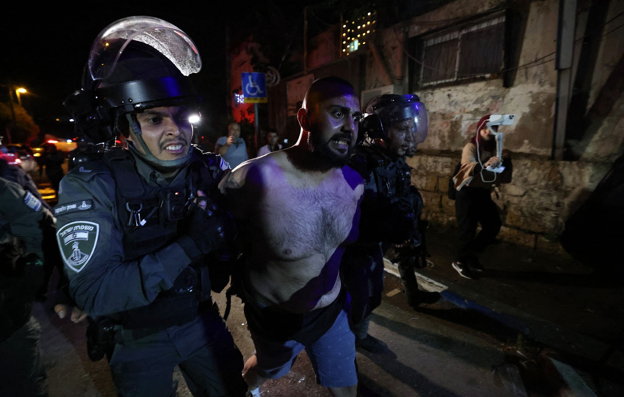 تظهر الصورة مجموعة من رجال الشرطة الذين يقومون باعتقال رجل. يبدو الرجل عاري الصدر ويظهر عليه علامات التوتر أو المقاومة. تحيط به عناصر من الشرطة يرتدون معدات حماية، ويبدو أنهم يتخذون إجراءات للسيطرة عليه. الخلفية توحي بأن المكان ربما كان شاهداً على مظاهر من التوتر أو الاضطراب، مع وجود إضاءة خافتة تشكل جوًا متوترًا.