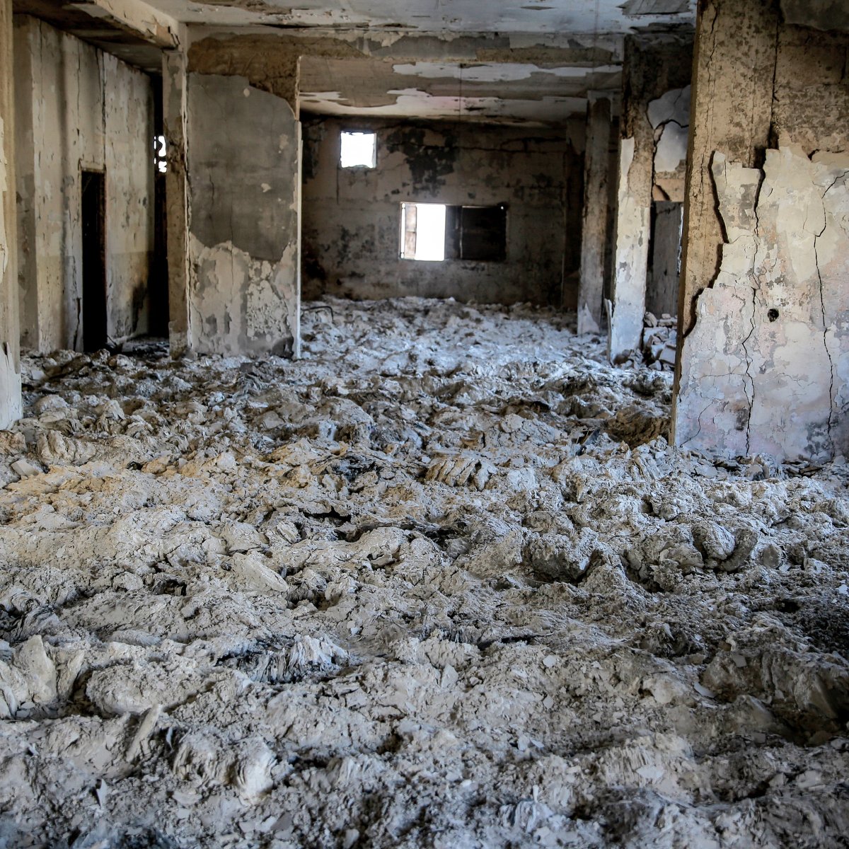 L'immagine mostra un interno di un edificio gravemente danneggiato. Le pareti sono deteriorate e presentano crepe, mentre il soffitto appare scrostato. Il pavimento è coperto da una massa bianca, che potrebbe essere polvere o detriti. I fori nelle pareti e nelle finestre suggeriscono che l'area è stata abbandonata e distrutta, creando un'atmosfera di desolazione e degrado. La luce che entra attraverso le aperture crea un contrasto con le ombre all'interno dello spazio.