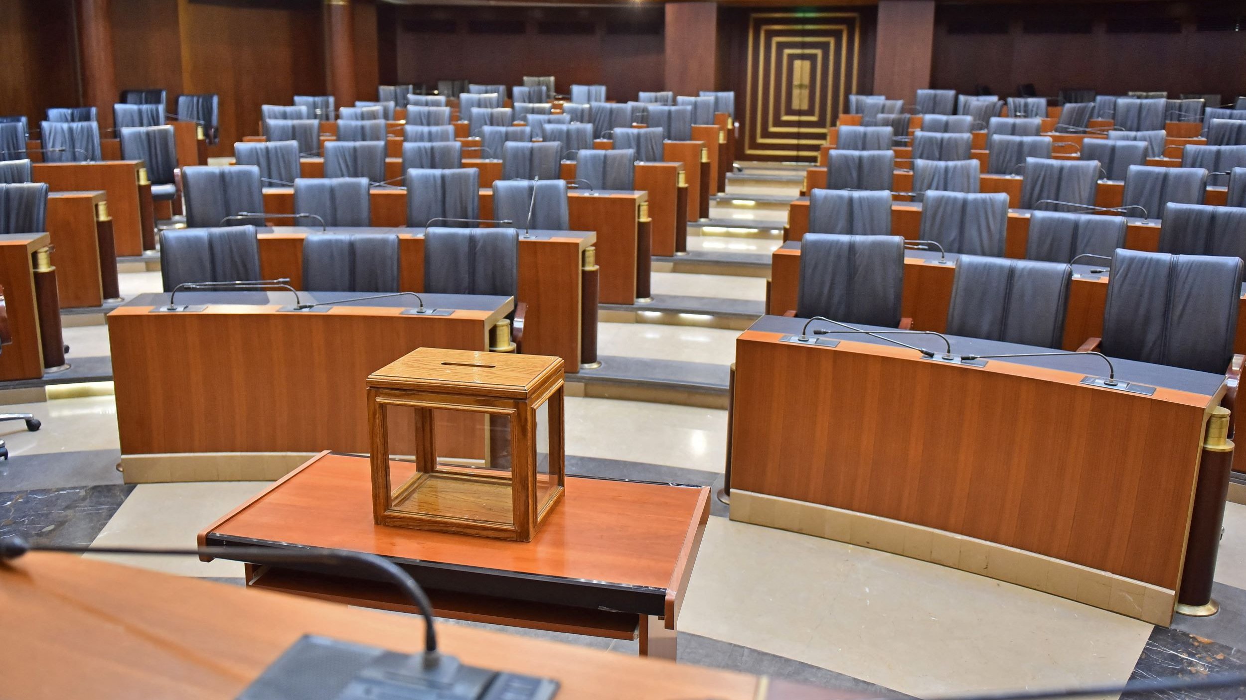 L'image montre une salle de réunion ou un parlement. Les sièges sont en bois, avec des dossiers noirs et sont disposés en rangées organisées. Au centre, il y a une petite table en bois avec une boîte en verre posée dessus. L'éclairage est doux, mettant en valeur les finitions en bois des murs et des meubles. L'ensemble de la salle dégage une atmosphère formelle et austère.