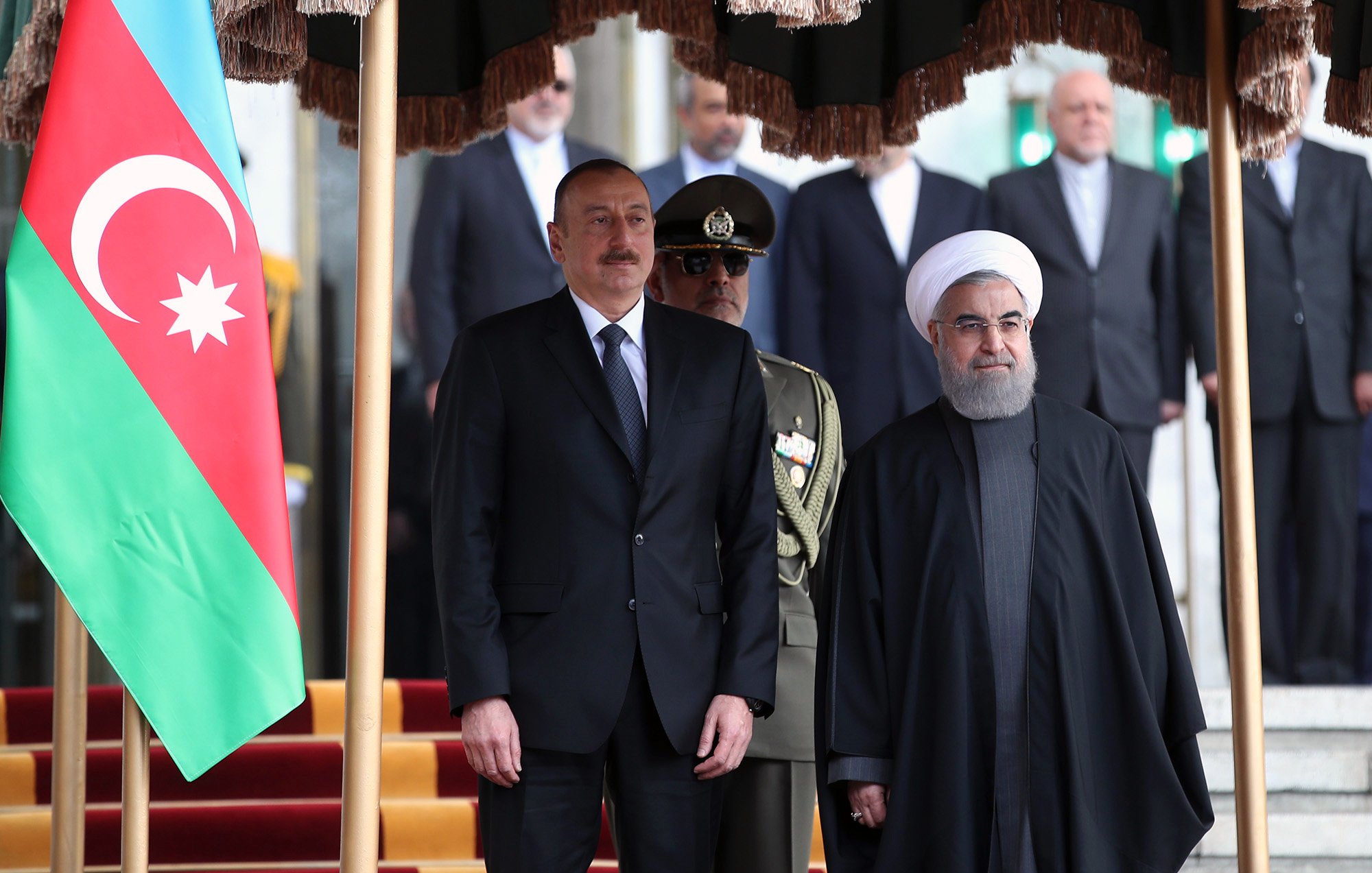 L'image montre deux hommes en costume, probablement des leaders politiques, posant ensemble devant un drapeau azéri. L'un d'eux porte un costume formel, tandis que l'autre est vêtu d'une tenue traditionnelle, avec une robe sombre. À l'arrière-plan, on aperçoit des soldats en uniforme et d'autres personnes, formant une sorte de cérémonie officielle. L'environnement semble être un bâtiment gouvernemental, avec des éléments décoratifs.