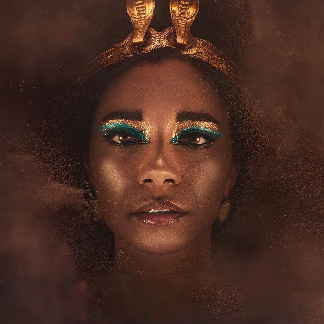 L'immagine presenta un volto femminile con un trucco molto elaborato, caratterizzato da ombretti blu e oro che mettono in risalto gli occhi. La persona indossa una corona decorata con serpenti dorati, che conferisce un aspetto regale e misterioso. Lo sfondo è sfocato e sembra essere avvolto da una polvere dorata, creando un'atmosfera affascinante e drammatica. La luce è calda e accentua la bellezza del viso, rendendo l'immagine suggestiva e intrigante.