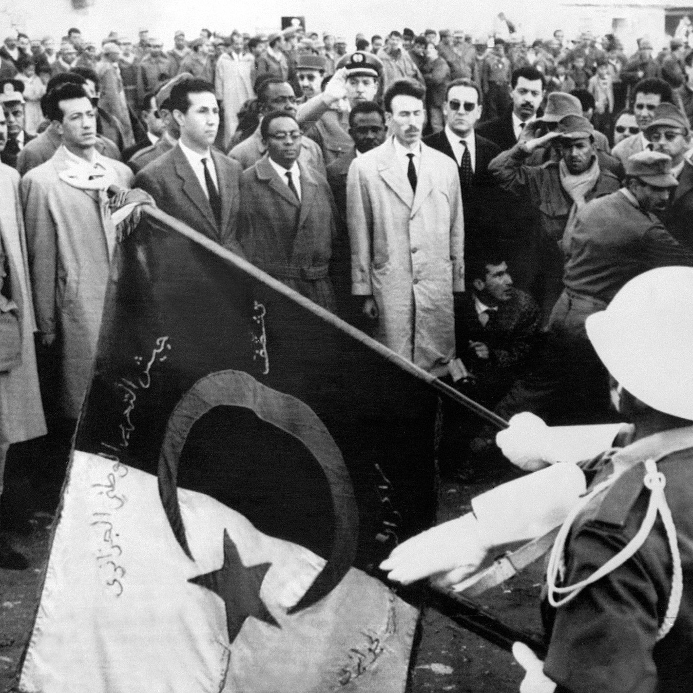 La imagen muestra un evento significativo, donde un grupo de personas se congrega. En el primer plano, soldados sostienen una bandera con símbolos distintivos, mientras que un grupo de hombres observa atentamente, algunos de ellos en posiciones de saludo. El ambiente parece ser solemne y formal, posiblemente relacionado con un momento histórico o ceremonioso. La vestimenta de los asistentes varía, con algunos en trajes y otros en uniformes militares, lo que sugiere una mezcla de civil y militar en el evento.