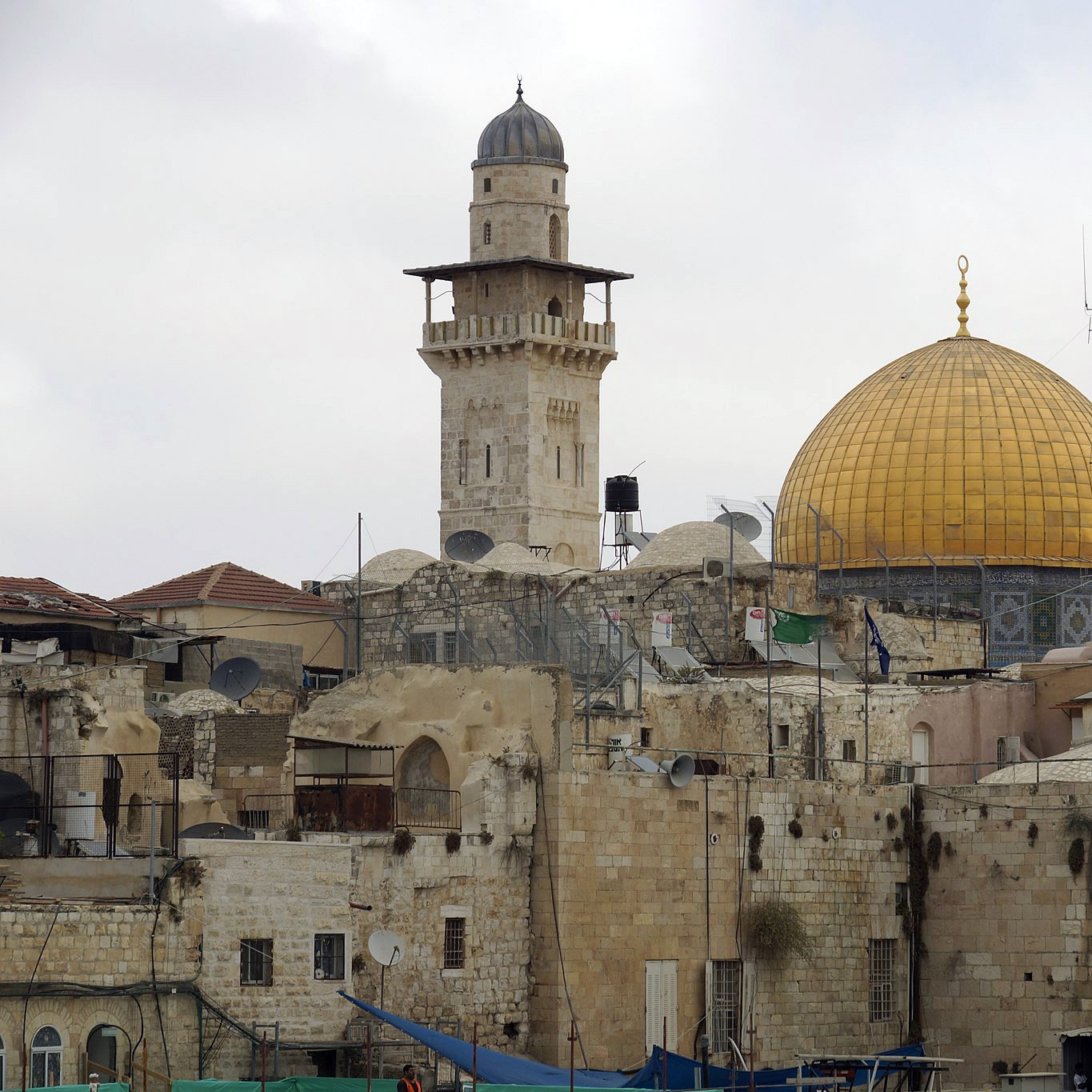 تظهر الصورة مشهداً لمدينة القدس، حيث يتوسطها المسجد الأقصى، الذي يبرز بقبة ذهبية مميزة. بجانب القبة، يمكن رؤية مئذنة ترتفع عالياً، وهناك بعض المباني التاريخية ذات الحجر القديم. كما يظهر في الصورة وجود أجهزة استقبال الأقمار الصناعية على أسطح المباني، مما يعكس مزيجاً من التاريخ والحداثة في هذه المنطقة. السماء تبدو غائمة، مما يضفي على المشهد جوّاً من السكون.
