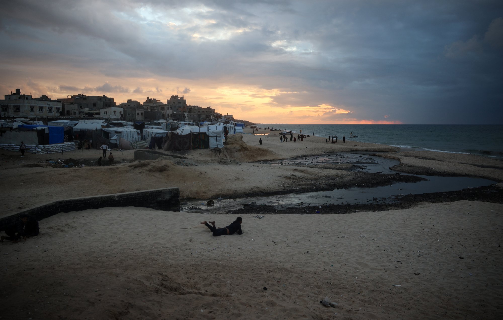 L'image montre une plage à l'heure du coucher de soleil, avec des nuages sombres dans le ciel. Au premier plan, on aperçoit du sable et une petite étendue d'eau stagnante. On peut également voir des tentes ou des abris, probablement des habitations temporaires, ainsi que quelques personnes se déplaçant sur la plage. L'atmosphère est à la fois paisible et mélancolique, avec des teintes orange et grises dans le ciel.