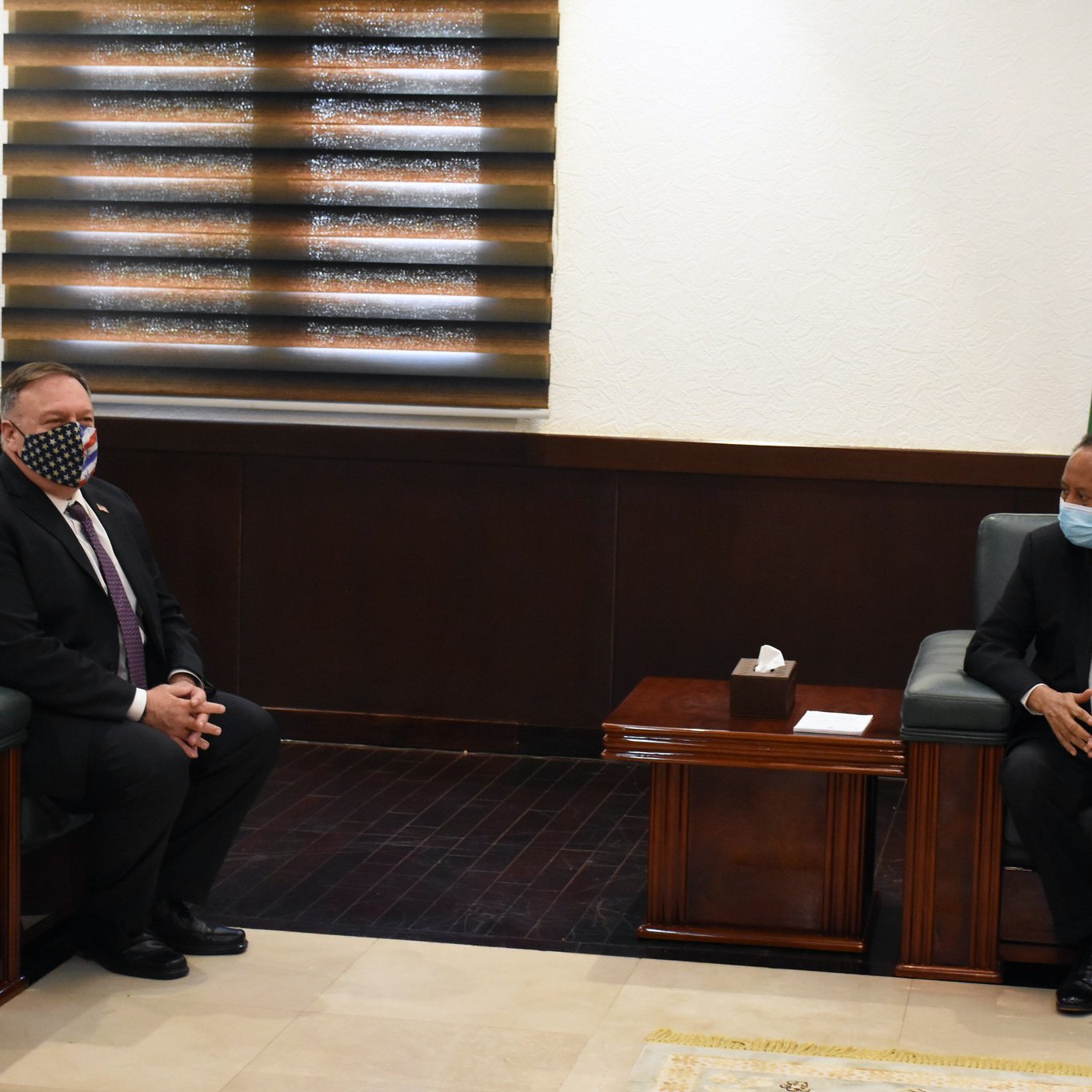 L'image montre deux hommes assis dans une pièce bien éclairée, avec des murs clairs et un sol en marbre. L'un des hommes porte un masque facial à motifs, tandis que l'autre est en costume avec une cravate. À l'arrière-plan, on peut voir un drapeau. Les deux hommes semblent engagés dans une conversation ou une réunion. Les meubles sont en bois sombre, et la pièce dégage une atmosphère formelle.