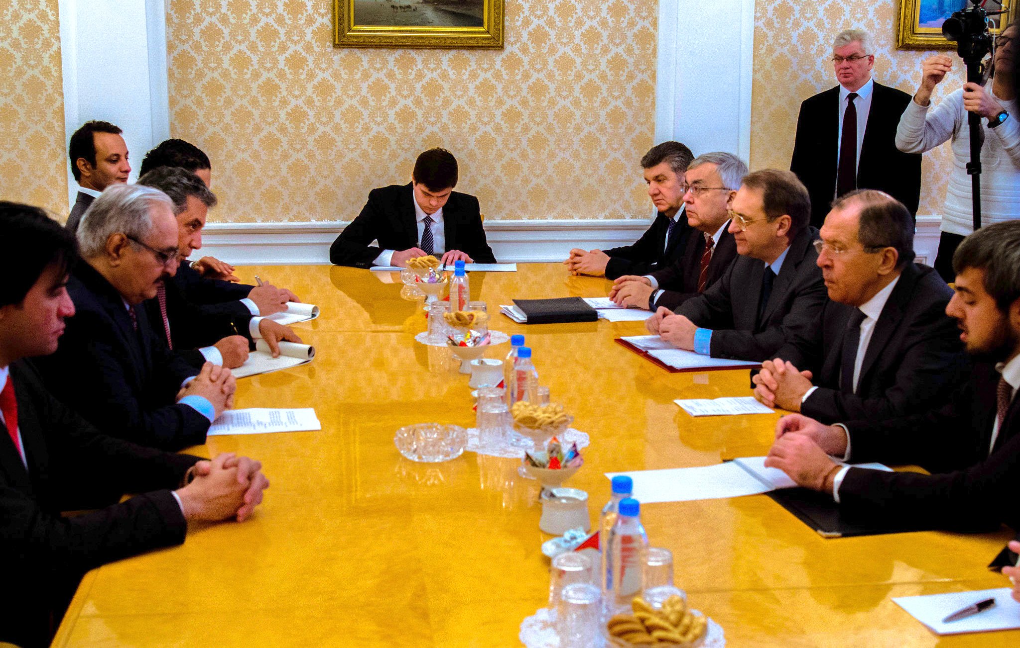 The image depicts a formal meeting taking place around a long wooden table in what appears to be a conference room. Participants are seated on both sides of the table, engaged in conversation or listening attentively. The table is set with various documents, and there are snacks and beverages in front of some attendees. The room has an elegant decor with a patterned wall and paintings. A few individuals are standing in the background, likely observing the meeting. The atmosphere seems serious and focused on discussion.