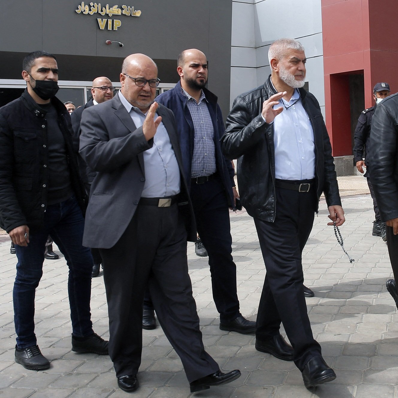 L'image montre un groupe de plusieurs hommes marchant ensemble à l'extérieur. Ils semblent se déplacer avec assurance et saluent peut-être des personnes présentes. Les hommes portent des vêtements variés, allant de vestes en cuir à des chemises et des pantalons sombres. L'environnement autour d'eux semble être un espace public, peut-être à proximité d'un bâtiment. Certains membres du groupe portent des masques. L'atmosphère paraît informelle mais organisée.