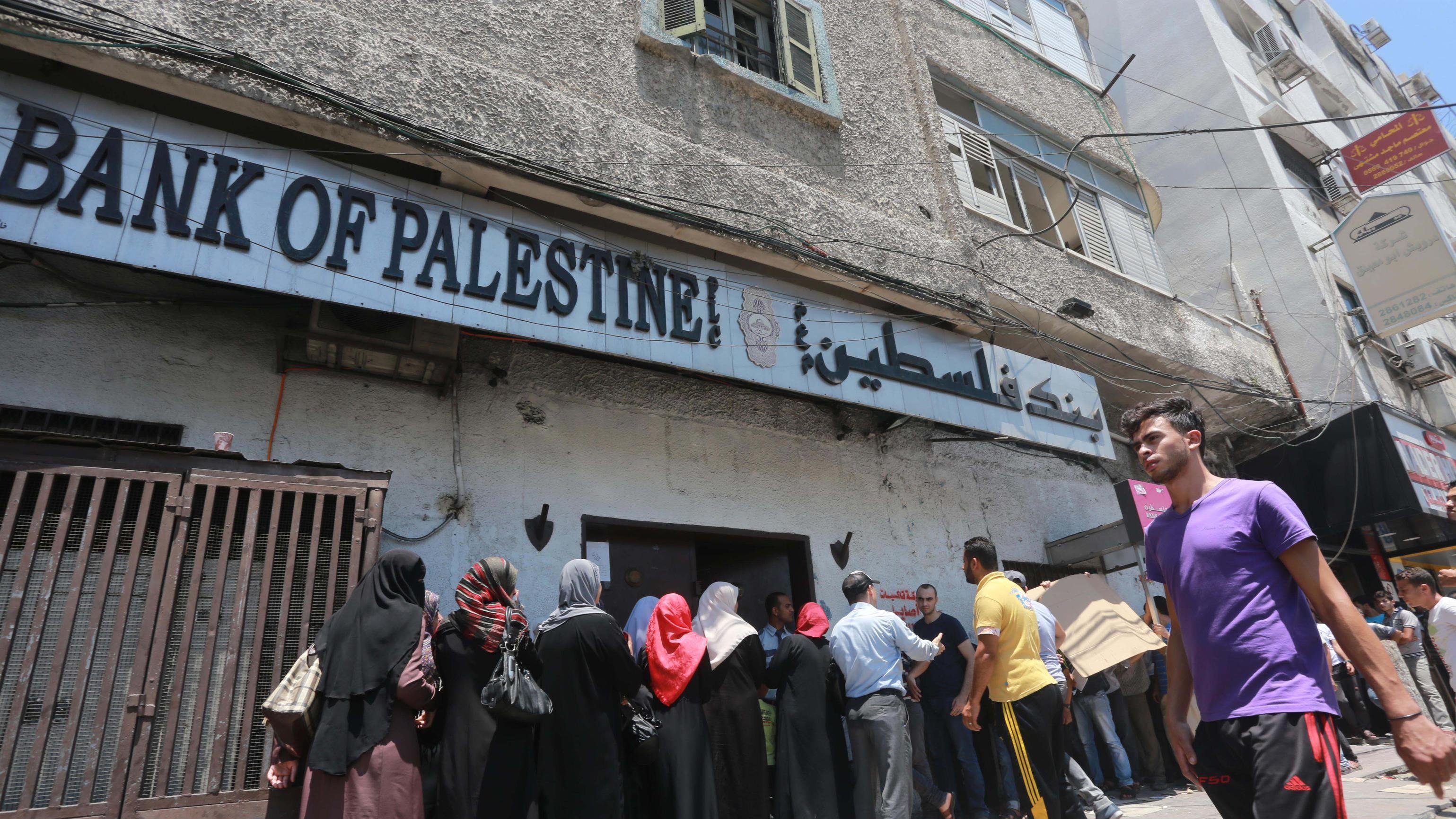 L'image montre une scène extérieure près d'un bâtiment portant l'inscription "Bank of Palestine". On y voit une file de personnes, principalement des femmes portant des vêtements traditionnels, attendant à l'extérieur de la banque. Certains hommes sont également présents, et on aperçoit un groupe d'individus qui se tiennent plus près de l'entrée. En arrière-plan, le bâtiment est ancien et les conditions semblent assez chaotiques, avec des câbles électriques visibles et des fenêtres en état d'usage. Le temps semble ensoleillé, suggérant une ambiance chaude.