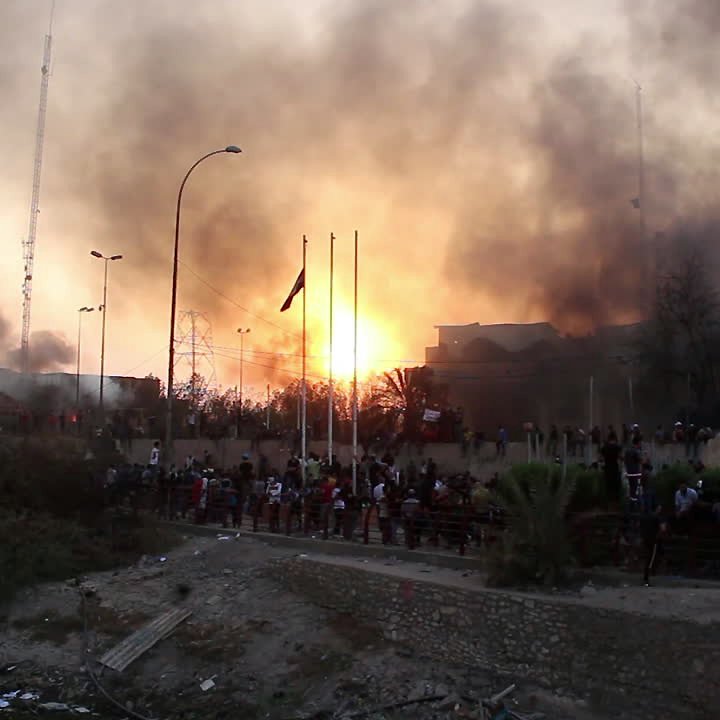 L'image montre une scène tumultueuse, probablement au cours d'un événement de protestation ou de conflit. On peut voir de la fumée s'élever dans le ciel, créant une atmosphère chaotique. À l'arrière-plan, un grand incendie semble se produire, projetant une lumière orange intense. Au premier plan, un groupe de personnes se rassemble, probablement en train d'observer ou de participer à l'événement. La présence de poteaux et de drapeaux suggère un espace urbain, peut-être une place publique ou un bord de fleuve. L'ensemble donne une impression d'urgence et de tension.
