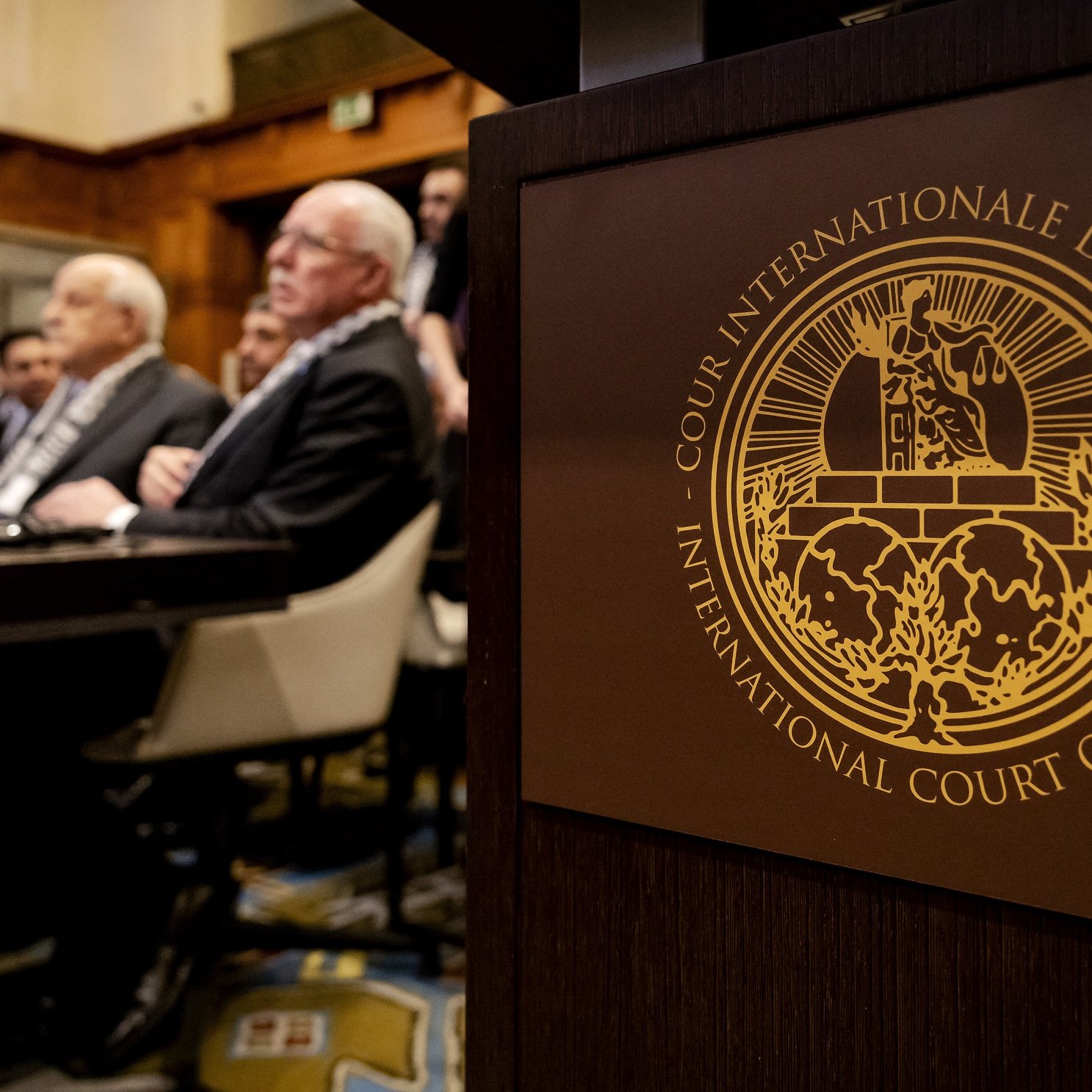 تظهر الصورة مشهدًا من داخل قاعة محكمة، حيث يجلس مجموعة من الأشخاص في الخلفية، ويبدو أنهم يستمعون لمداولات أو حديث مهم. في المقدمة، يوجد شعار المحكمة الدولية للعدل، وهو يتضمن تصميمًا معقدًا يرمز إلى العدالة. الطراز المعماري للقاعة يظهر الفخامة والرسمية. الأجواء تعكس أهمية الحدث الحاصل داخل هذه المحكمة.