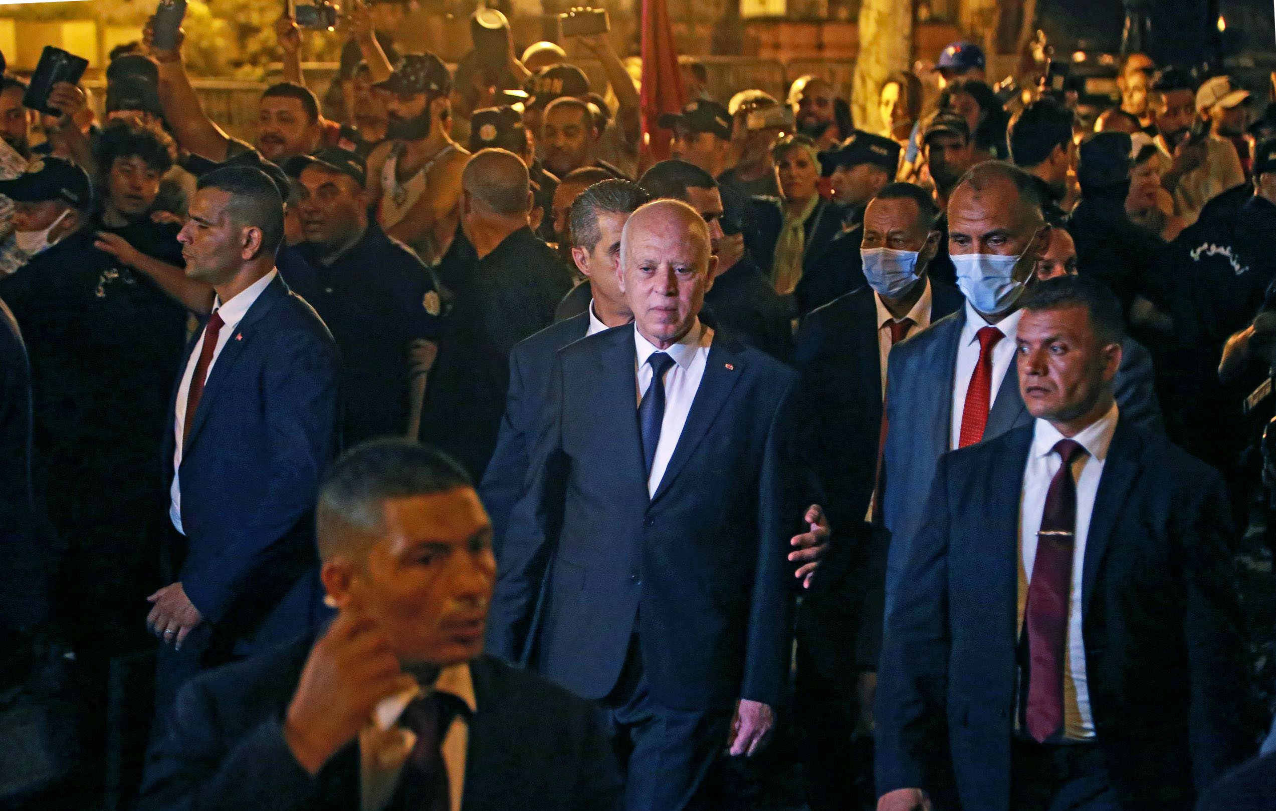 L'image montre un groupe de personnes rassemblées autour d'un homme en costume noir, qui semble être une figure politique ou publique. Il est entouré de plusieurs membres de ce qui semble être une équipe de sécurité, tous vêtus de costumes sombres. Les gens en arrière-plan semblent attentifs et certains utilisent des téléphones pour prendre des photos. L'ambiance paraît animée, avec un éclairage qui suggère qu'il fait sombre.