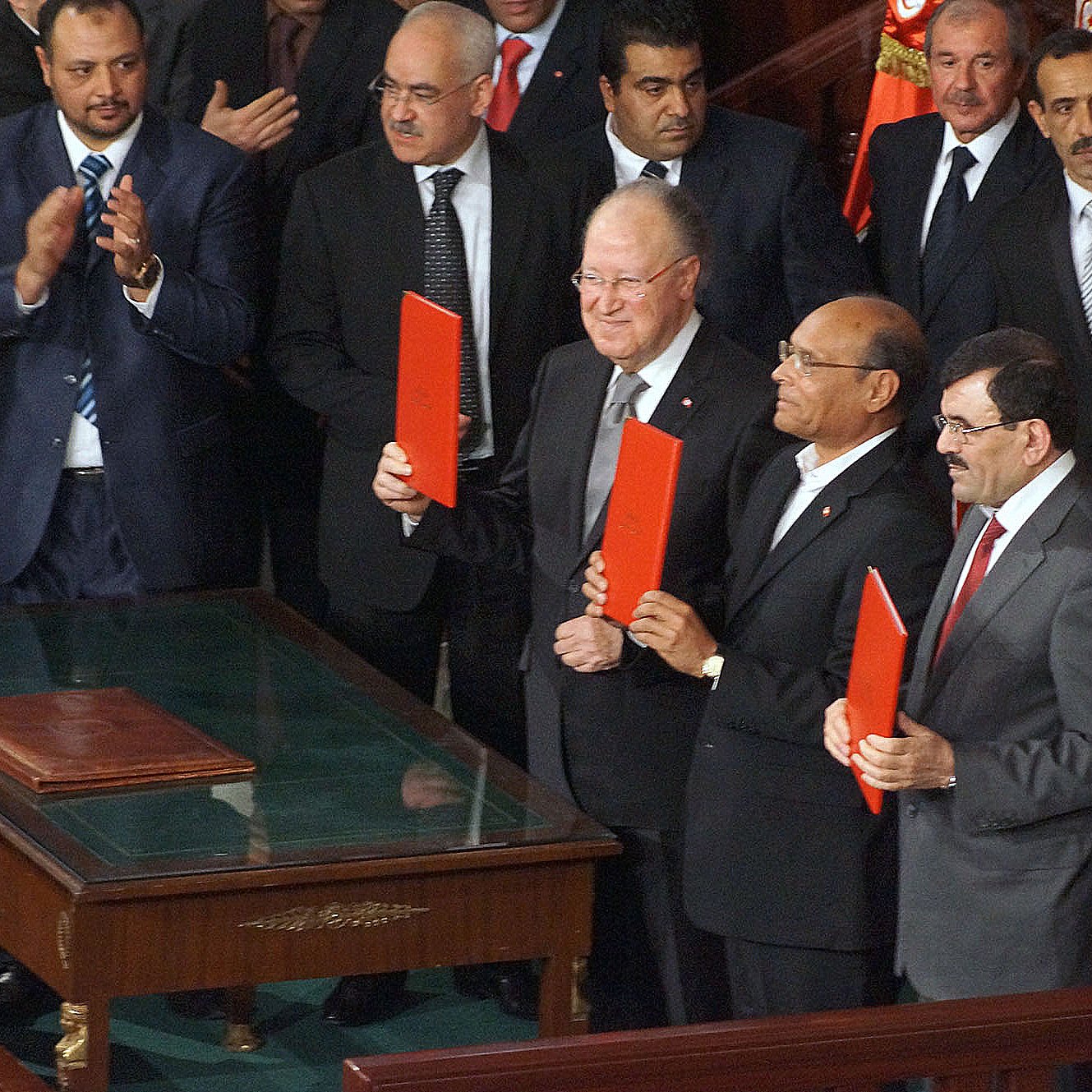 L'image montre un groupe d'hommes se tenant debout autour d'une table, tous souriants et tenant des dossiers rouges. Ils semblent être dans un cadre officiel, probablement lors d'une cérémonie ou d'une réunion importante. En arrière-plan, on peut voir d'autres personnes assises et applaudissant, ainsi que des drapeaux. L'ambiance semble solennelle et festive à la fois.
