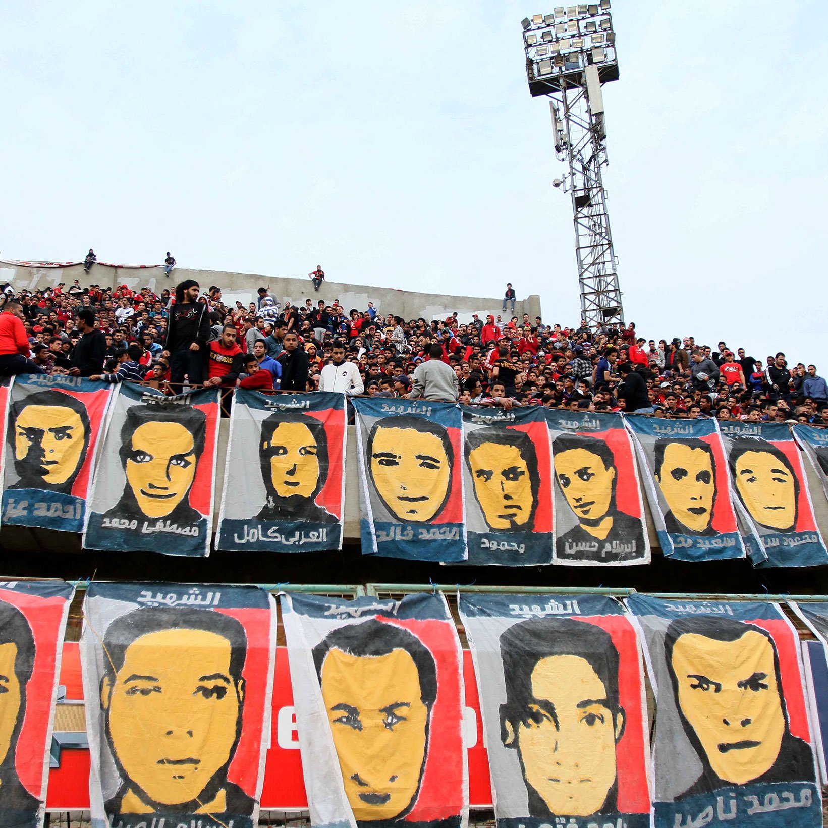 L'image montre un grand groupe de supporters dans un stade, rassemblés dans les gradins. Ils sont entourés de nombreuses banderoles affichant des portraits de différentes personnes, probablement des joueurs ou des figures importantes liés à l'équipe. L'ambiance semble festive et engagée, avec les fans montrant leur passion pour leur équipe. Le ciel est nuageux et le stade est bien rempli de spectateurs.