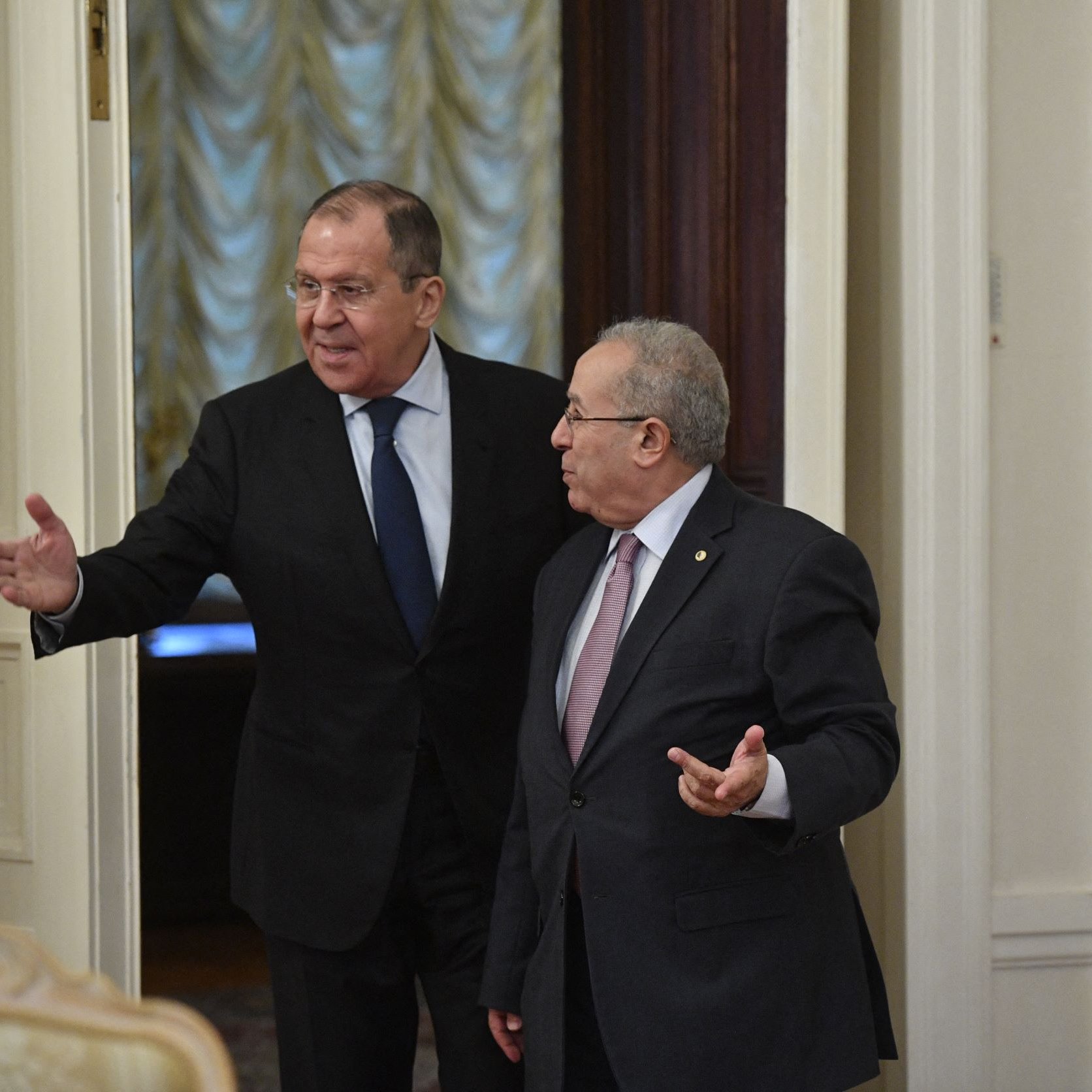 The image shows two men in a formal setting, likely inside a government building. One man is gesturing with his hand as he walks, indicating he is speaking or inviting the other man to follow. The second man appears to be listening attentively. Both are dressed in formal attire, suggesting a diplomatic or official meeting. The room has a sophisticated decor, with elements such as elegant walls and furniture visible in the background.