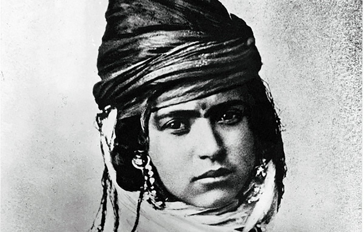 L'image montre un portrait noir et blanc d'une personne portant un turban orné et des bijoux. Le regard de la personne est intense, et son expression transmet une certaine dignité. Elle est vêtue de vêtements traditionnels qui reflètent une culture spécifique. Les détails du turban et des ornements ajoutent une dimension riche et texturée à l'image.