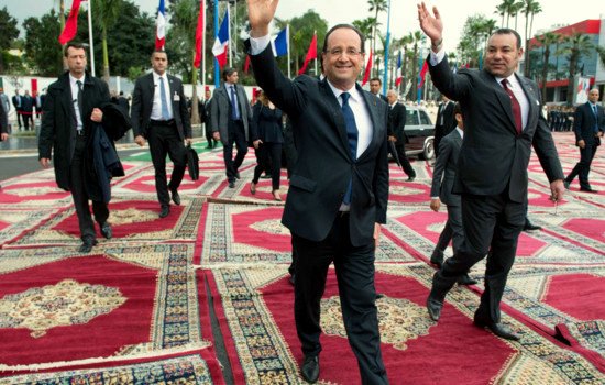 L'image montre deux hommes marchant sur un tapis richement décoré. L'un d'eux, vêtu d'un costume sombre et souriant, fait un geste de la main, tandis que l'autre, également en costume, marche à ses côtés. En arrière-plan, on peut voir des drapeaux de différentes nations, ce qui suggère un événement international ou officiel. L'atmosphère semble conviviale et solennelle à la fois.