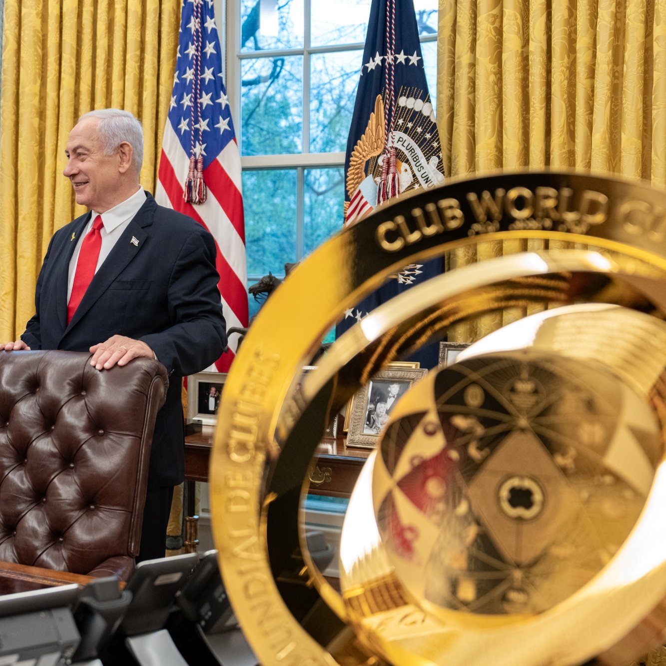 Two men in suits stand in an ornate office with gold curtains and flags, one holding a small object.