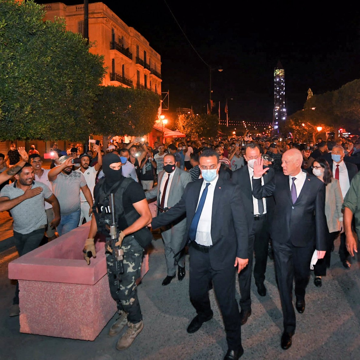 La imagen muestra a un grupo de personas en una calle durante la noche. En el centro, hay un hombre vestido con traje y corbata, escoltado por un guardia de seguridad con una máscara y uniforme. Alrededor, se ven varios manifestantes levantando las manos, algunos parecen estar enérgicos o en protesta. La iluminación de la escena proviene de faroles y luces en el entorno, creando un ambiente animado y posiblemente tenso. Los árboles y edificios en el fondo sugieren que se trata de una zona urbana.