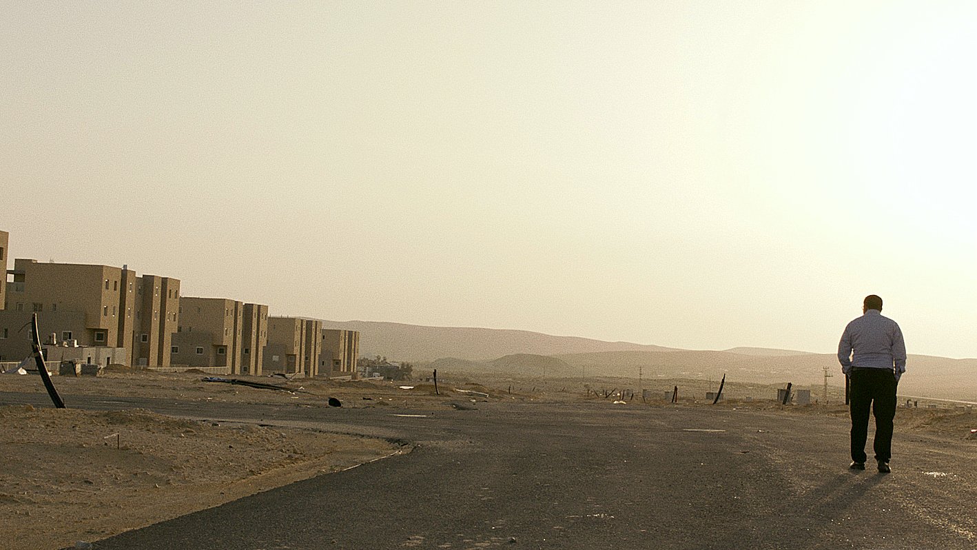 L'image montre un homme marchant seul sur une route déserte. On peut voir en arrière-plan des bâtiments modernes qui semblent inachevés ou abandonnés, avec un paysage désertique s'étendant à l'horizon. La lumière du soleil semble douce, suggérant une ambiance calme et un peu mélancolique. Le ciel est clair, et l'ensemble de la scène évoque une sensation de solitude et d'isolement.