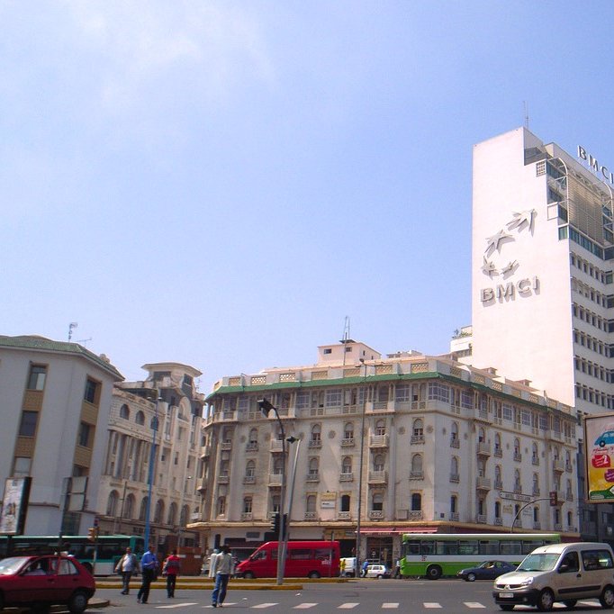 The image shows a cityscape featuring a tall building with the letters "BMCI" prominently displayed on it. The architecture of the surrounding buildings is a mix of modern and traditional styles. The sky is clear and blue, indicating a sunny day. In the foreground, there are vehicles on the road, including a red car and a few buses, as well as a billboard advertising various products. The scene conveys a bustling urban environment.