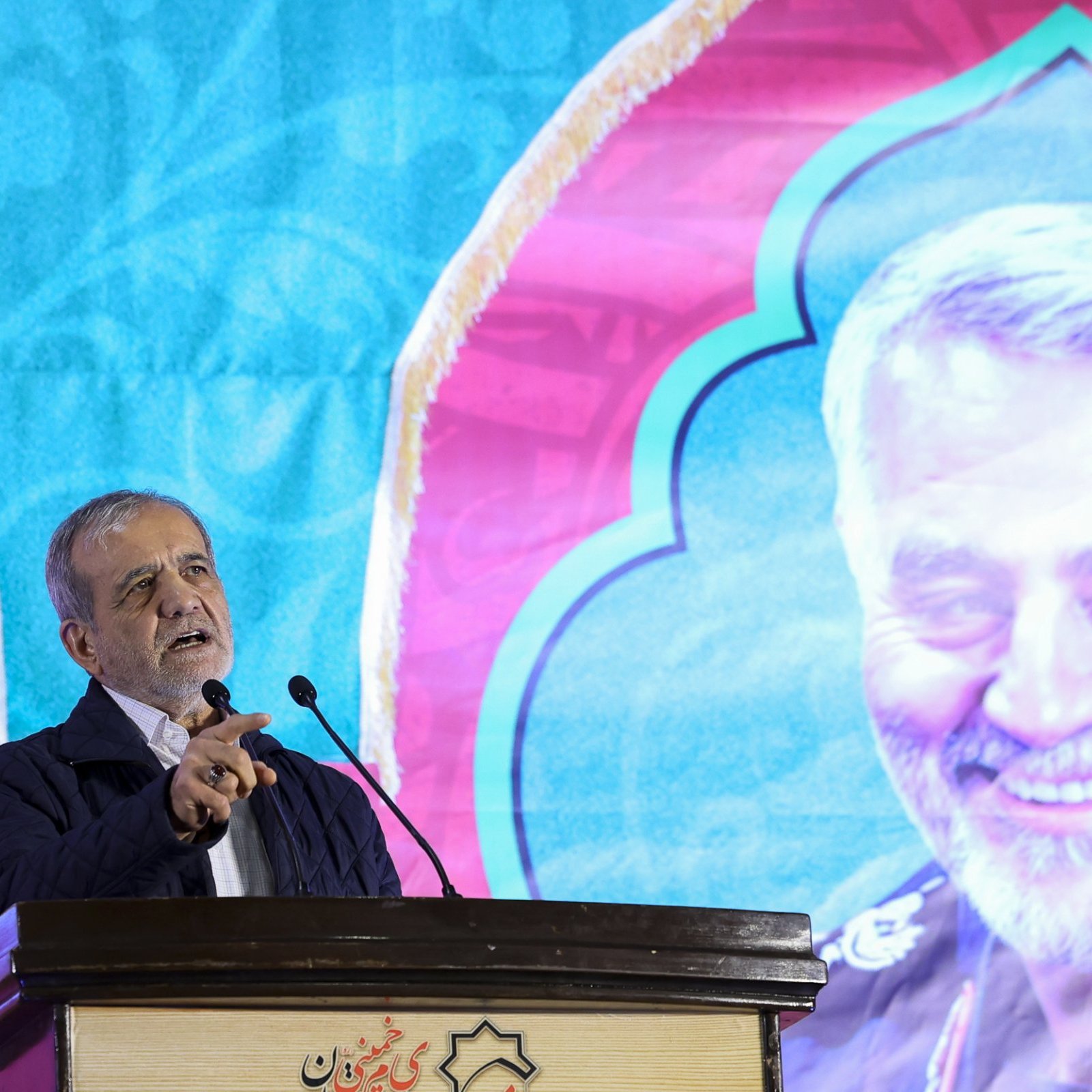 L'image montre un homme en train de s'exprimer lors d'un discours devant un microphone. En arrière-plan, il y a un grand panneau coloré affichant le portrait d'un homme souriant, probablement une figure importante. Le fond est décoré avec des motifs vibrants. L'homme au premier plan semble prendre la parole avec passion, pointant du doigt, ce qui indique qu'il parle probablement d'un sujet important ou émotionnel.