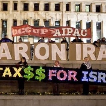 L'image montre un groupe de personnes réunies autour d'une grande pancarte ou d'un affichage, exprimant des revendications politiques. Ils tiennent des panneaux où l'on peut lire "NO WAR ON IRAN" et "NO US TAX$$ FOR ISRAEL". L'ambiance semble être celle d'une manifestation ou d'un rassemblement en faveur de la paix et critiquant l'utilisation des fonds publics. L'arrière-plan comprend un bâtiment, suggérant que l'action a lieu dans un espace public.