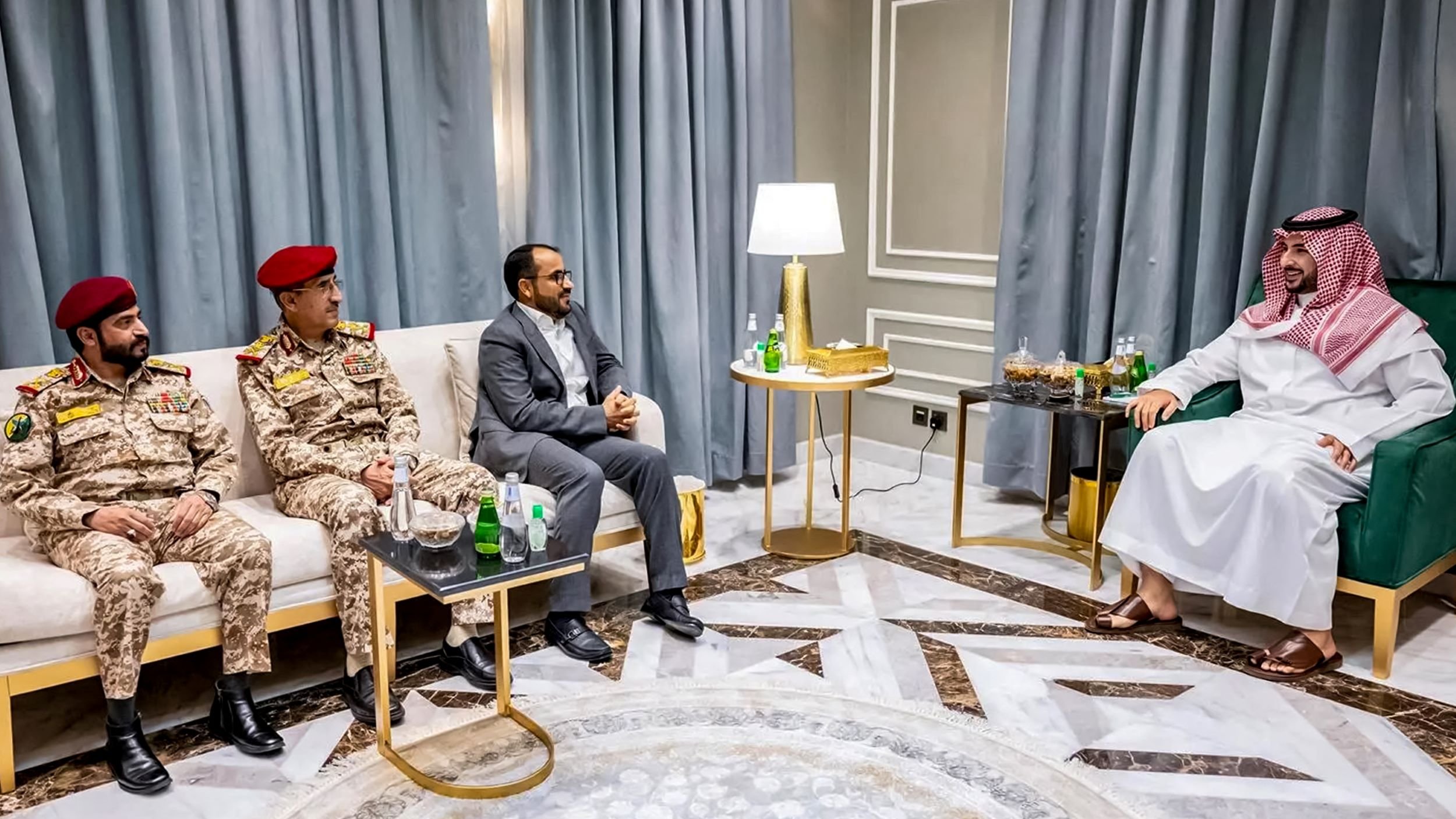 تظهر الصورة مجموعة من الأشخاص الذين يجلسون في غرفة معيشة أنيقة. في الجهة اليسرى، يوجد ضابطان عسكريان يرتديان زيًا عسكريًا، أحدهما يرتدي قبعة حمراء. بينما يجلس رجل ثالث في المنتصف بزي مدني. على اليمين، يوجد رجل يرتدي لباسًا تقليديًا سعوديًا (ثوب وغطاء رأس) يجلس على أريكة. يوجد أيضًا طاولة صغيرة أمامهم تحتوي على بعض المشروبات. الجدران مكسوة بستائر تتناسب مع الديكور الداخلي.