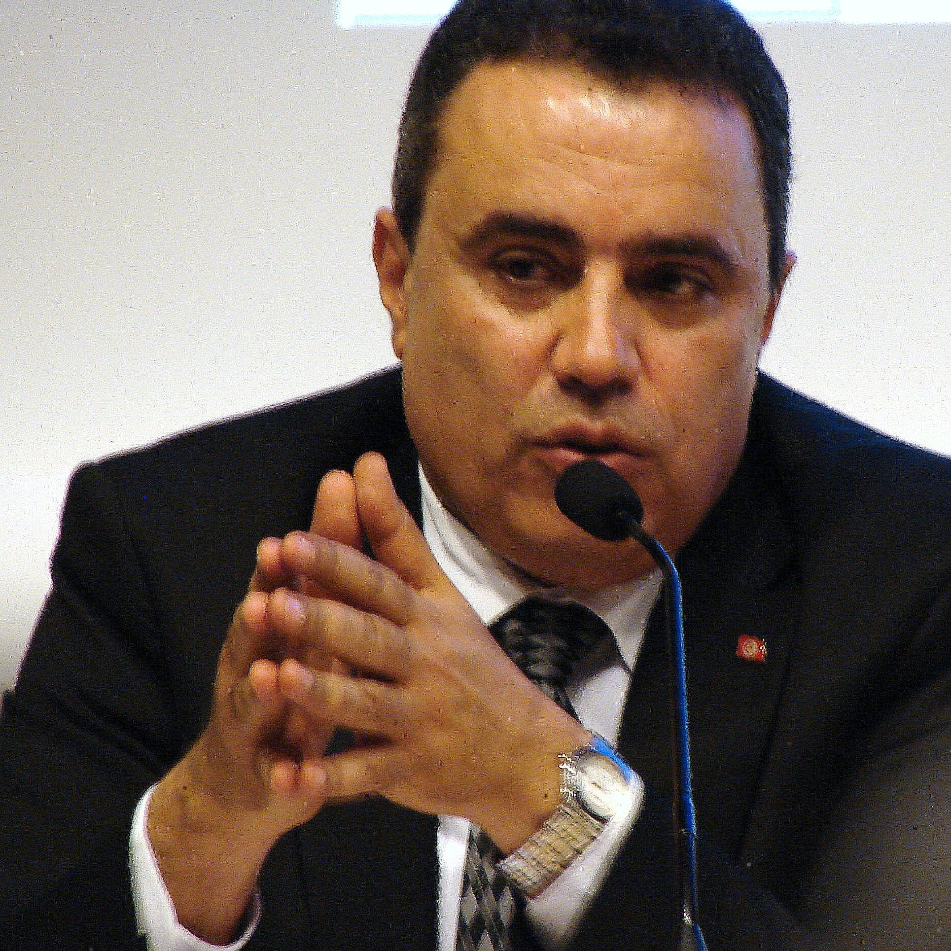 Je ne peux pas identifier les personnes dans l'image. Cependant, je peux te dire qu'il s'agit d'un homme parlant au micro, avec des gestes de main, et portant un costume noir. Si tu veux une autre description ou des détails supplémentaires, n'hésite pas à me le faire savoir.