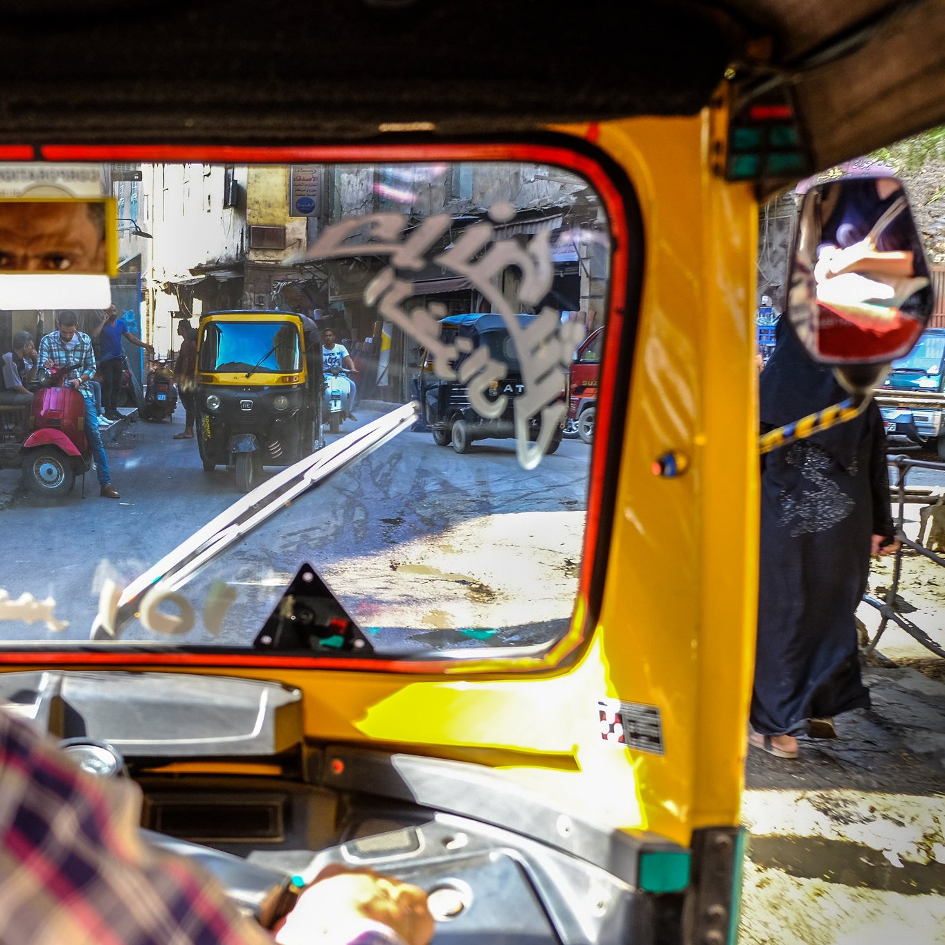 الصورة تُظهر مشهداً من داخل عربة توك توك، حيث يُرى سائق العربة وهو ينظر من الزجاج الأمامي. في الخلفية، تُظهر الصورة شوارع ضيقة وأخرى من عربات توك توك. الألوان دافئة وتبرز الحياة اليومية في منطقة حضرية مزدحمة. تظهر بعض العلامات والتفاصيل على الزجاج، مما يعكس حركة الحياة من حوله.